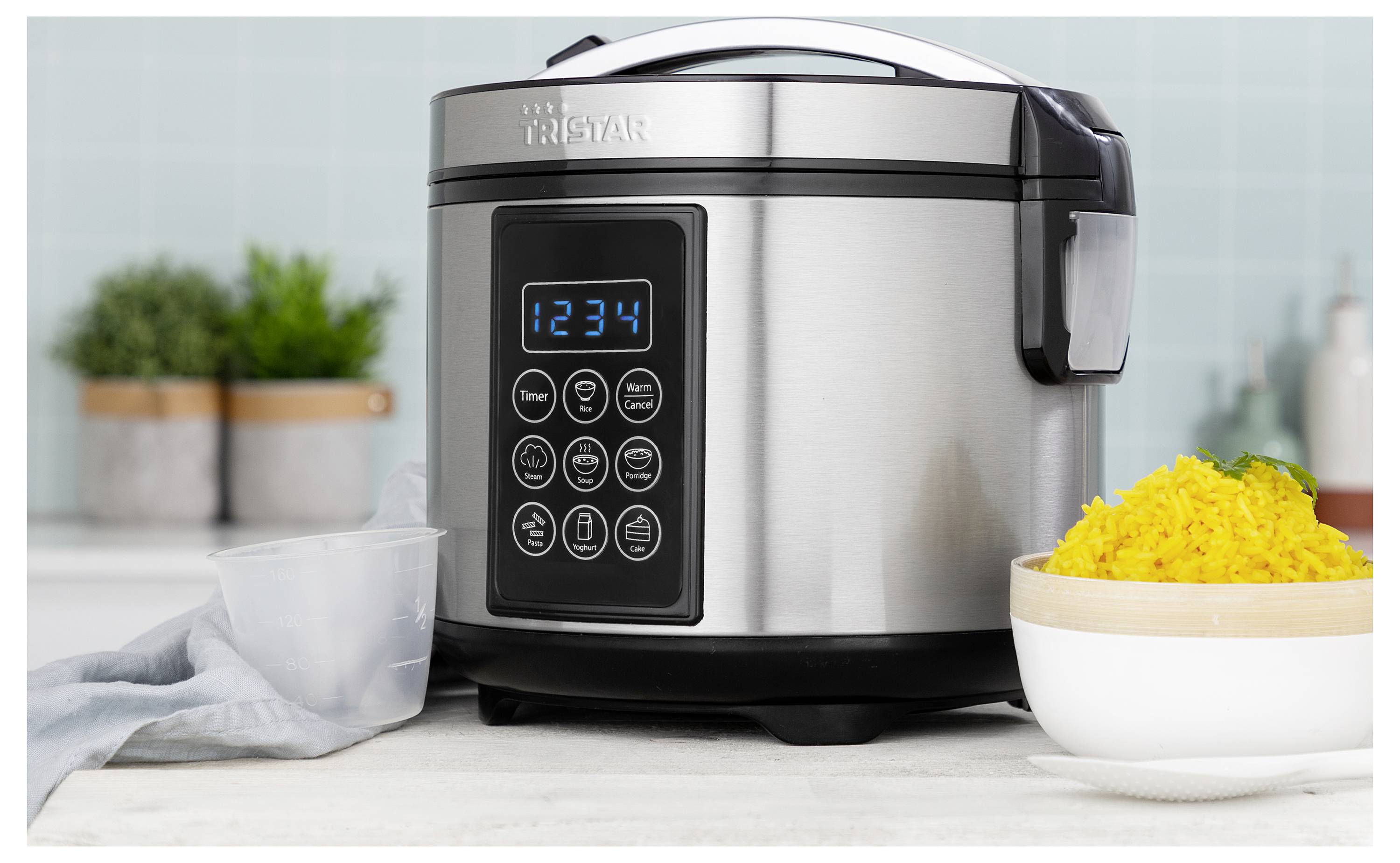 Stainless steel rice cooker with digital display showing '1234', placed beside a bowl of cooked yellow rice and measuring cup on a kitchen counter.