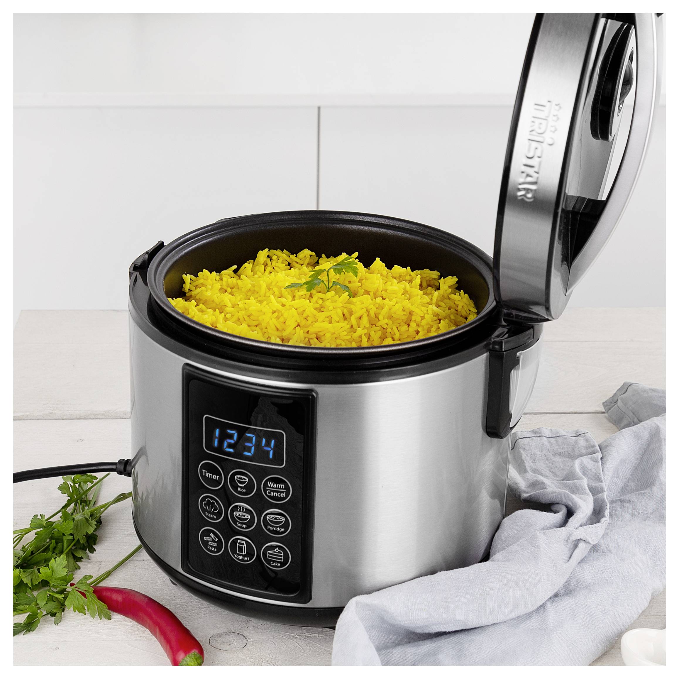 Open rice cooker filled with yellow rice, with digital controls visible, on a kitchen counter beside herbs, a chili pepper, and a cloth.