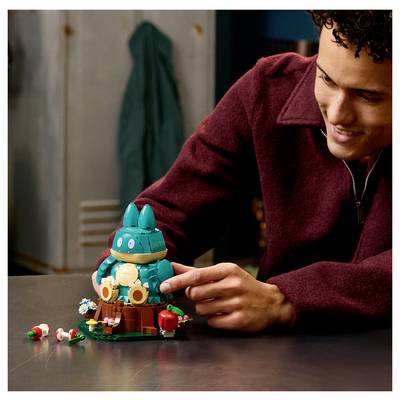 A man smiles while assembling a teal-and-yellow LEGO-style bunny figure with small flowers and accessories on a table.