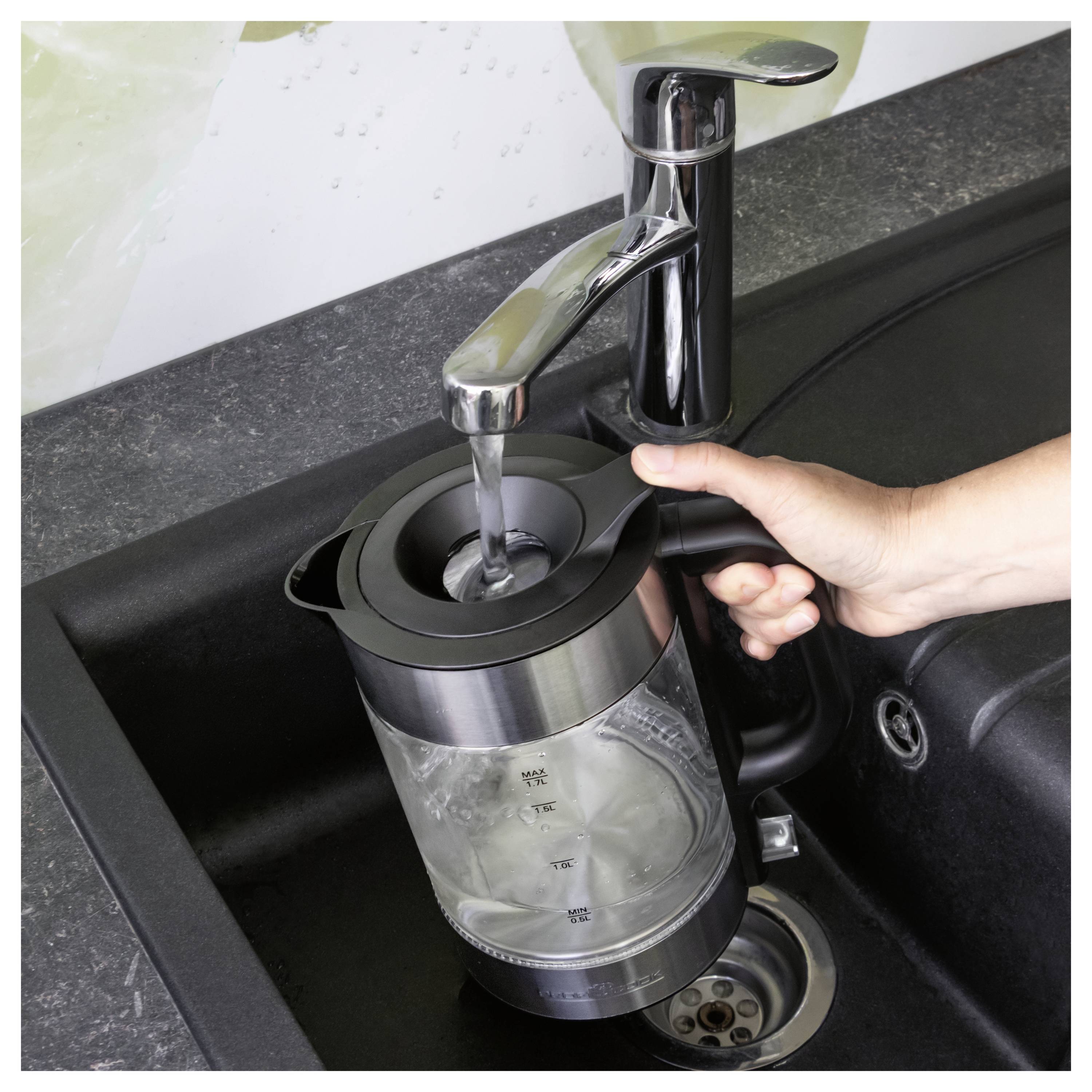 Hand rinsing a clear electric kettle under a kitchen faucet in a black sink.