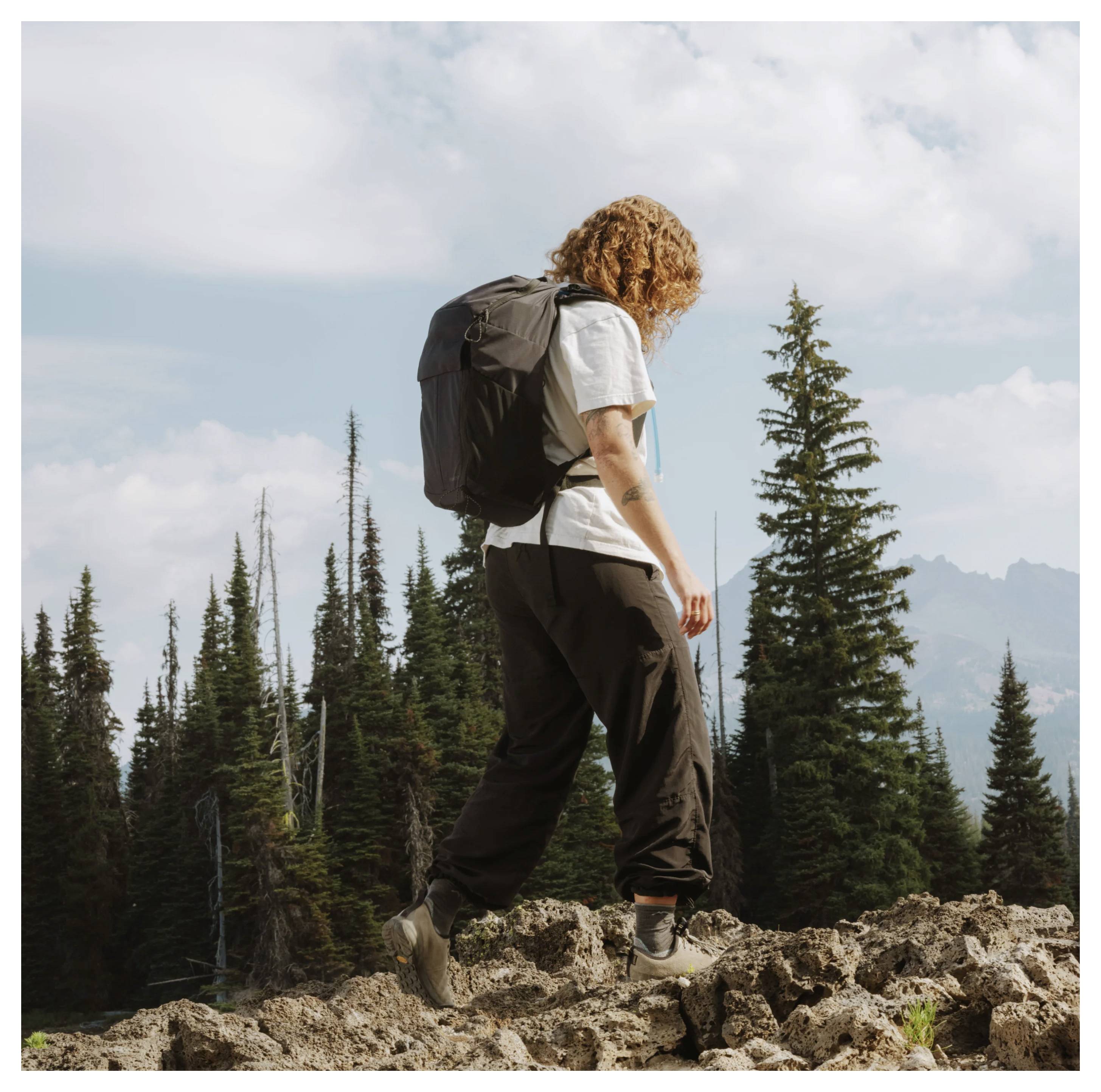 Hiker with a backpack walking across rocky terrain in a forested mountain area under a cloudy sky.