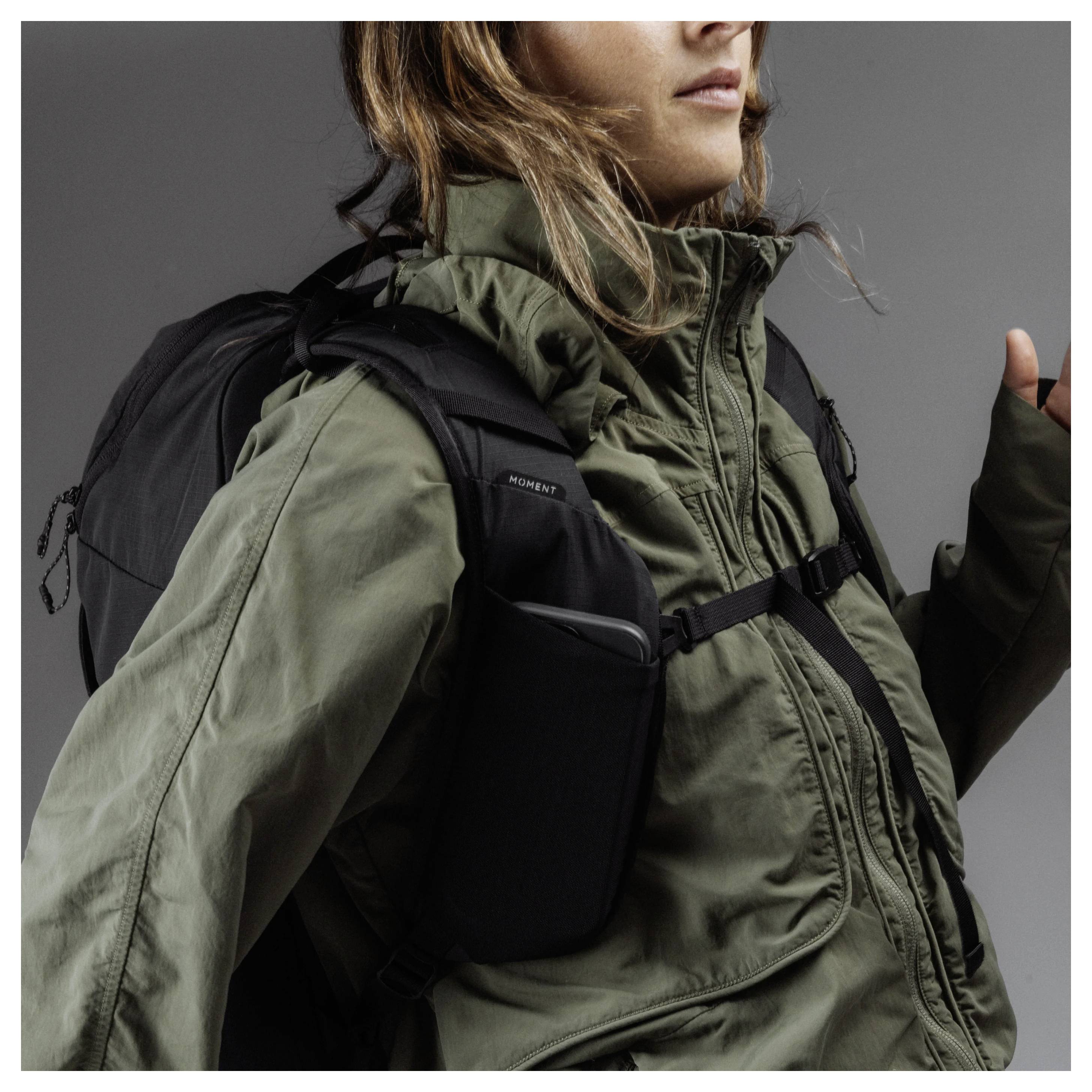 Woman wearing an olive green hooded jacket and black backpack against a gray studio background.