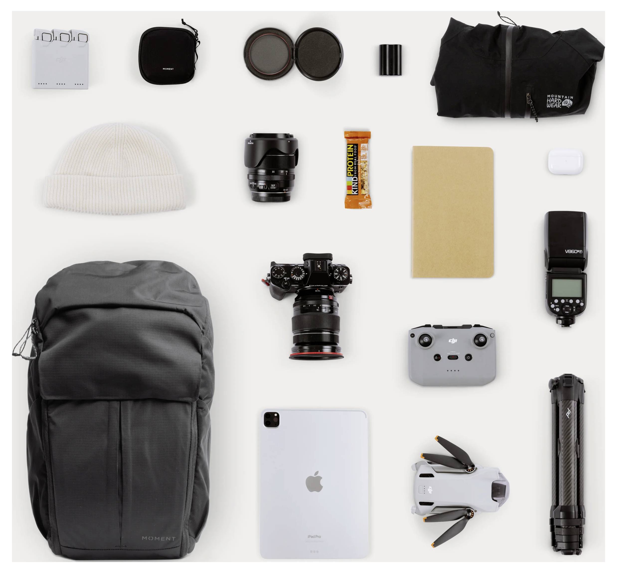 Flat lay of travel and photography gear: backpack, camera with lens, drone, iPad, flash, batteries, filters, beanie, notebook, tripod, and accessories arranged on a white background.
