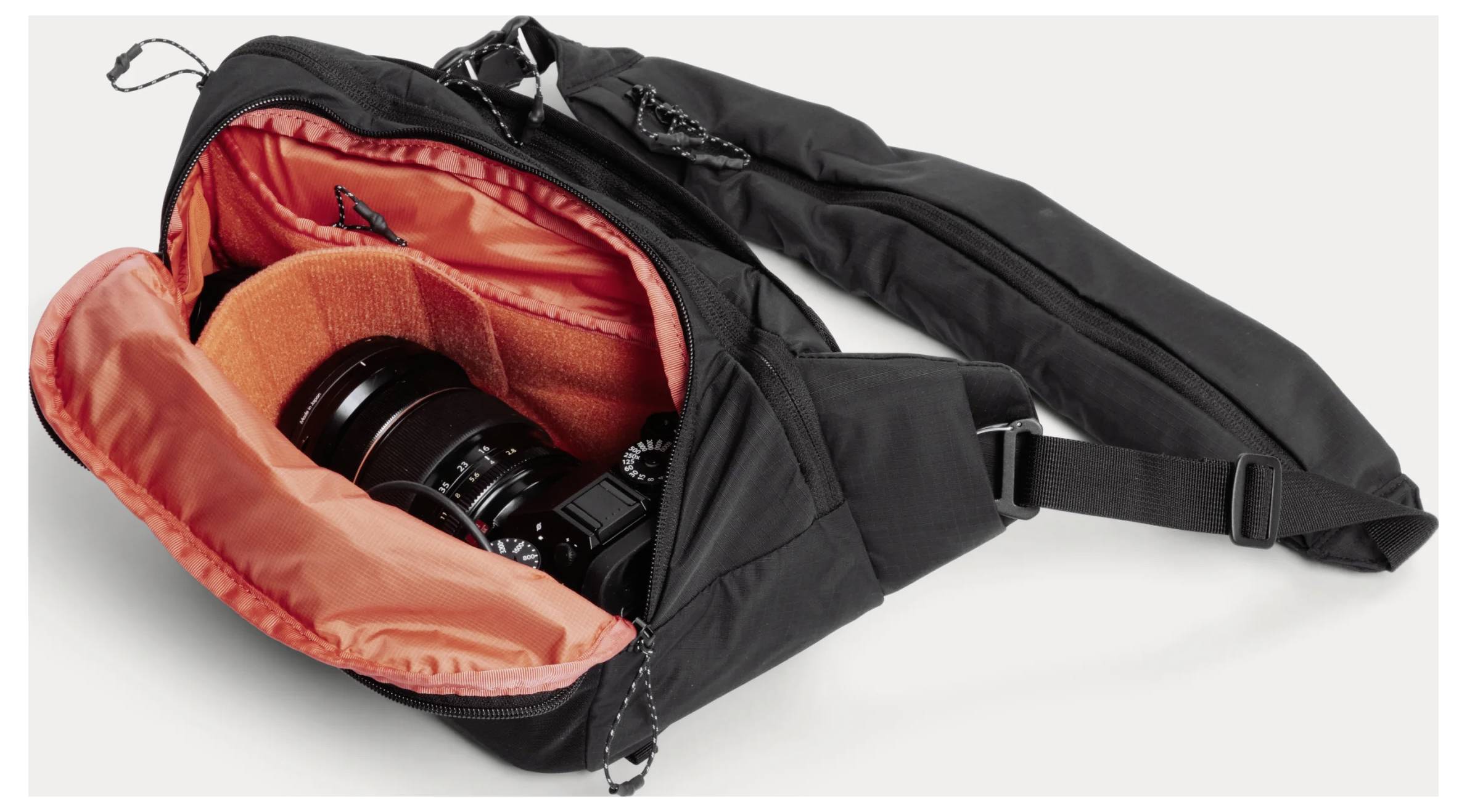 Black camera bag open to reveal orange padded interior with a camera lens and gear inside.