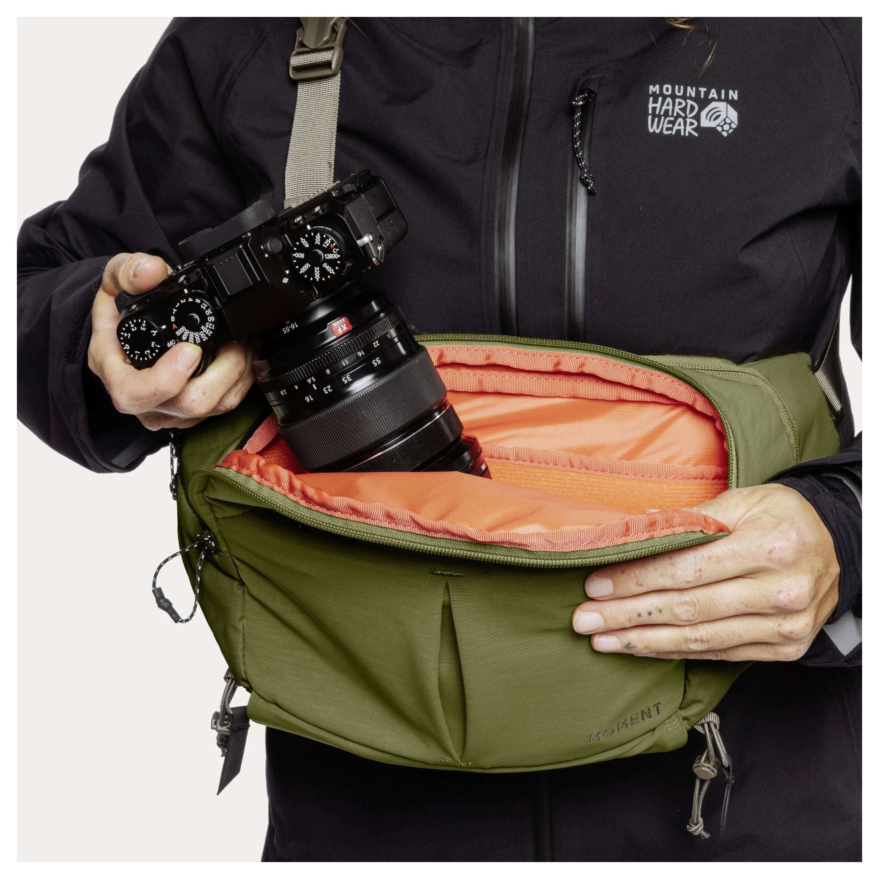 Person removing a camera with attached lens from an olive green waist pack with orange padded lining, showing gear storage use.