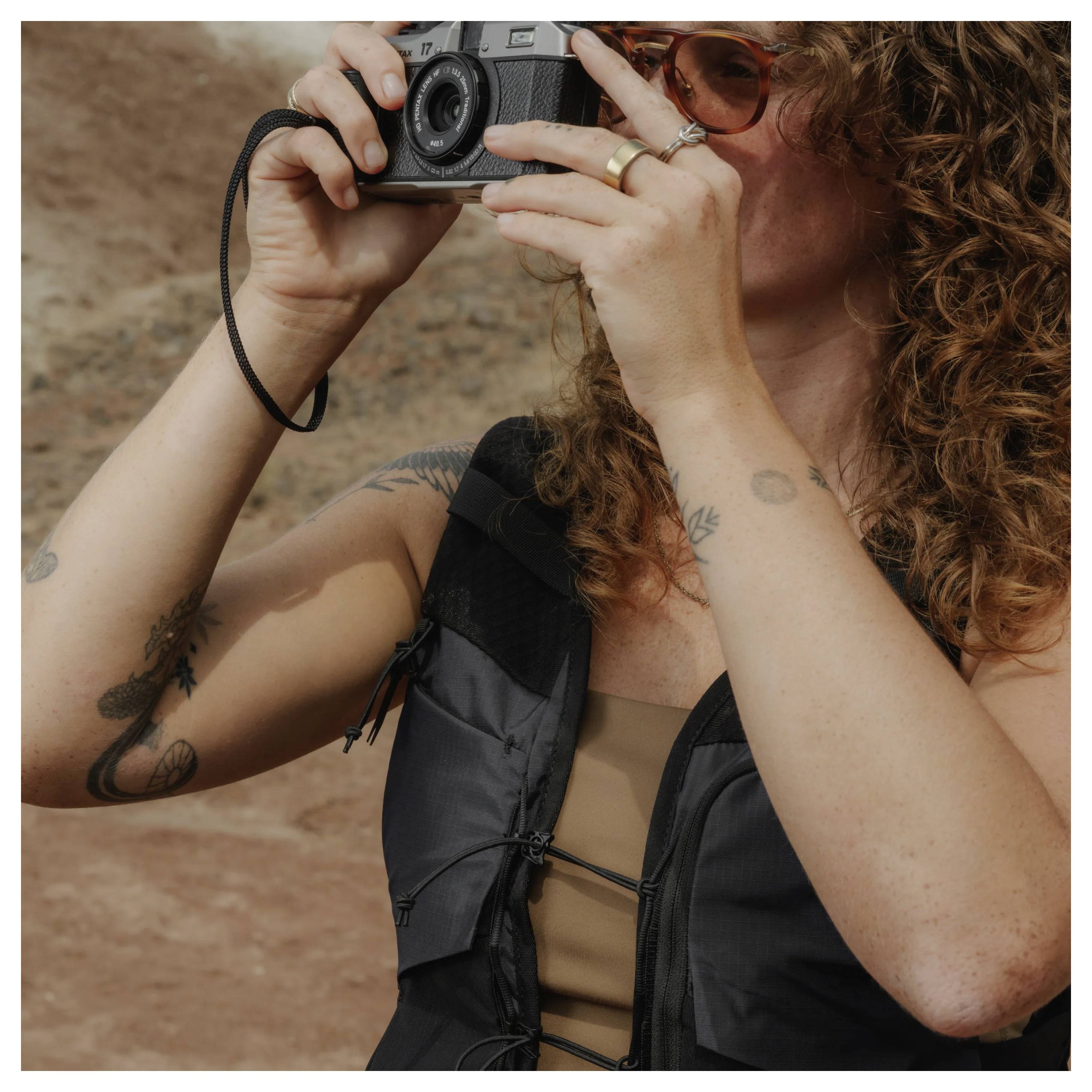 A woman with curly hair and sunglasses takes a photo with a camera, wearing a black top against a rocky outdoor background.