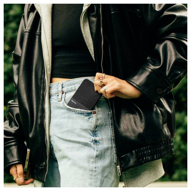 Person in black leather jacket and jeans holding a black smartphone partly pulled from front pocket.