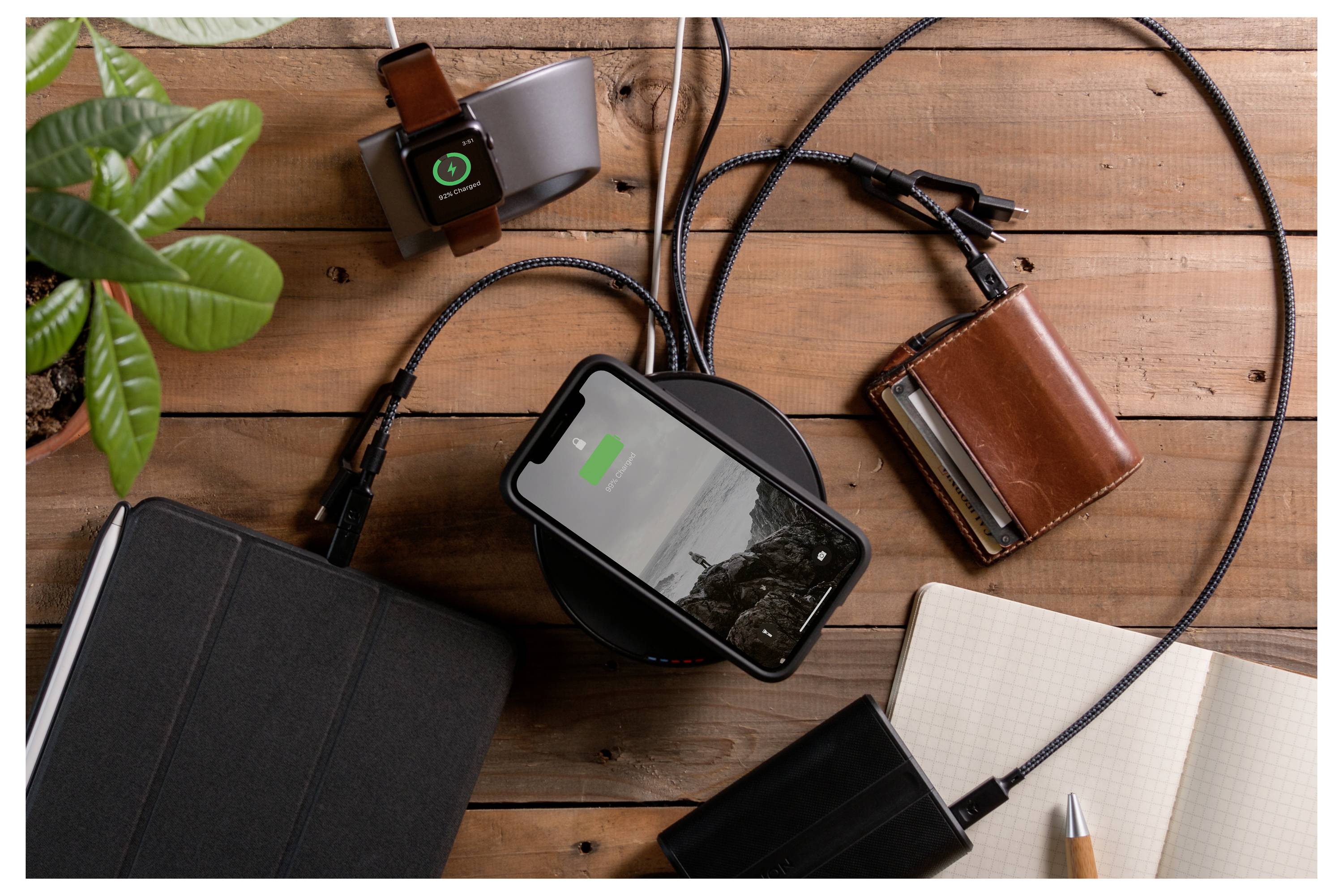 Wireless charger charging a smartphone on a wooden desk, surrounded by a smartwatch, notebook, wallet, cables, and a plant.