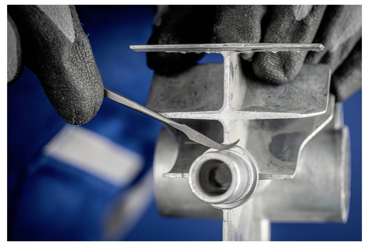 Gloved hands inspect a metal mechanical part with a pointed tool in a workshop setting.