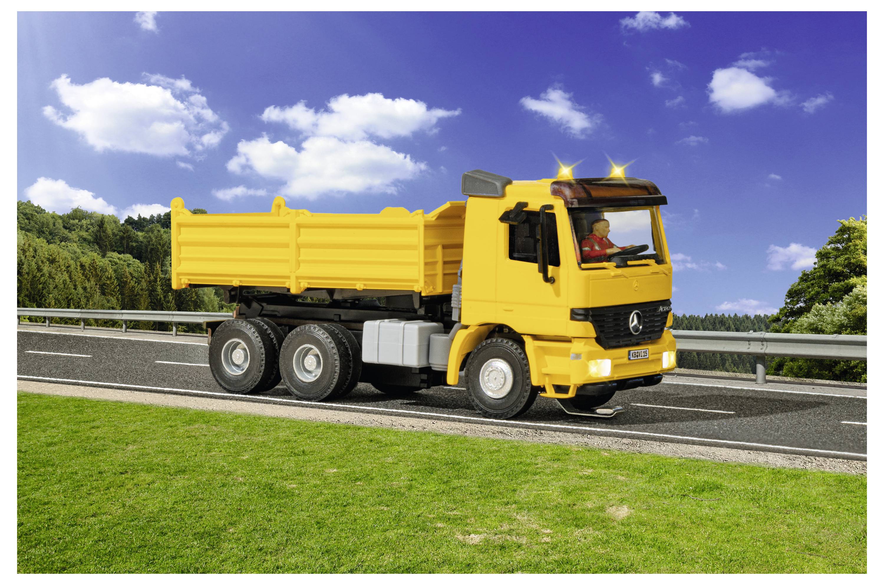 Yellow dump truck driving on a road beside grass and guardrails under a blue sky with clouds.