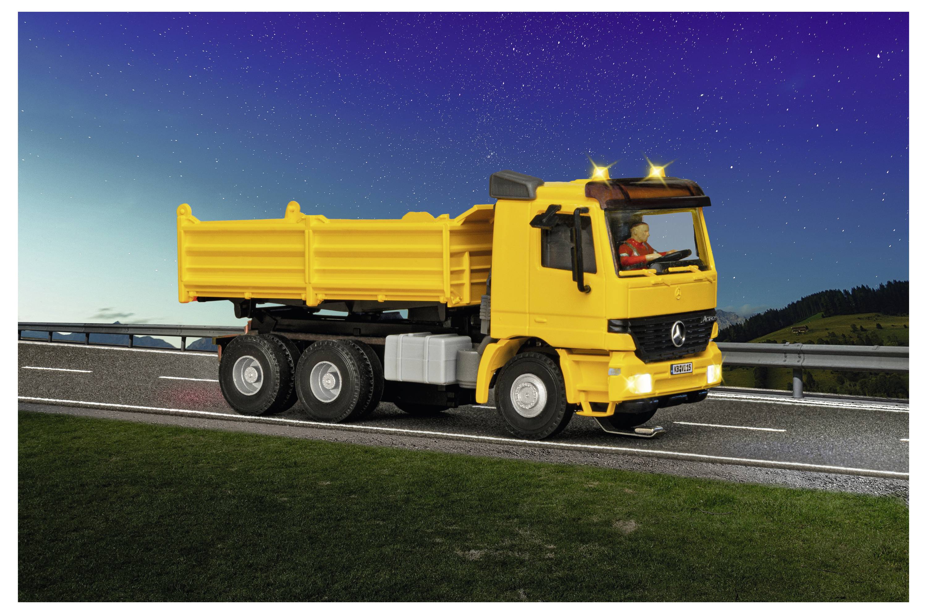 Bright yellow dump truck driving on a highway at night, with headlights on and a driver visible in the cab.