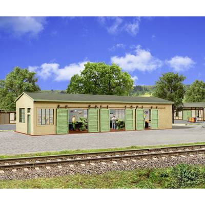 Model train station platform with a small beige building and green doors, workers inside, railroad tracks in the foreground, trees and blue sky behind.
