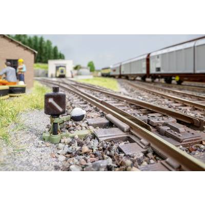 Close-up of a model railway switch beside tracks, with a freight train in the background and miniature workers near a trackside building.
