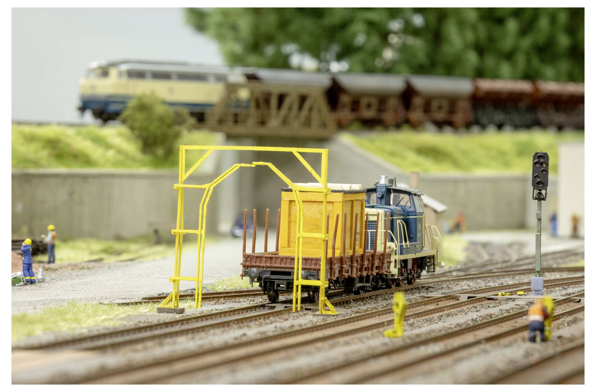 Model train scene with a blue locomotive pulling freight cars through a trackside maintenance yard, with miniature workers and a yellow gantry frame.