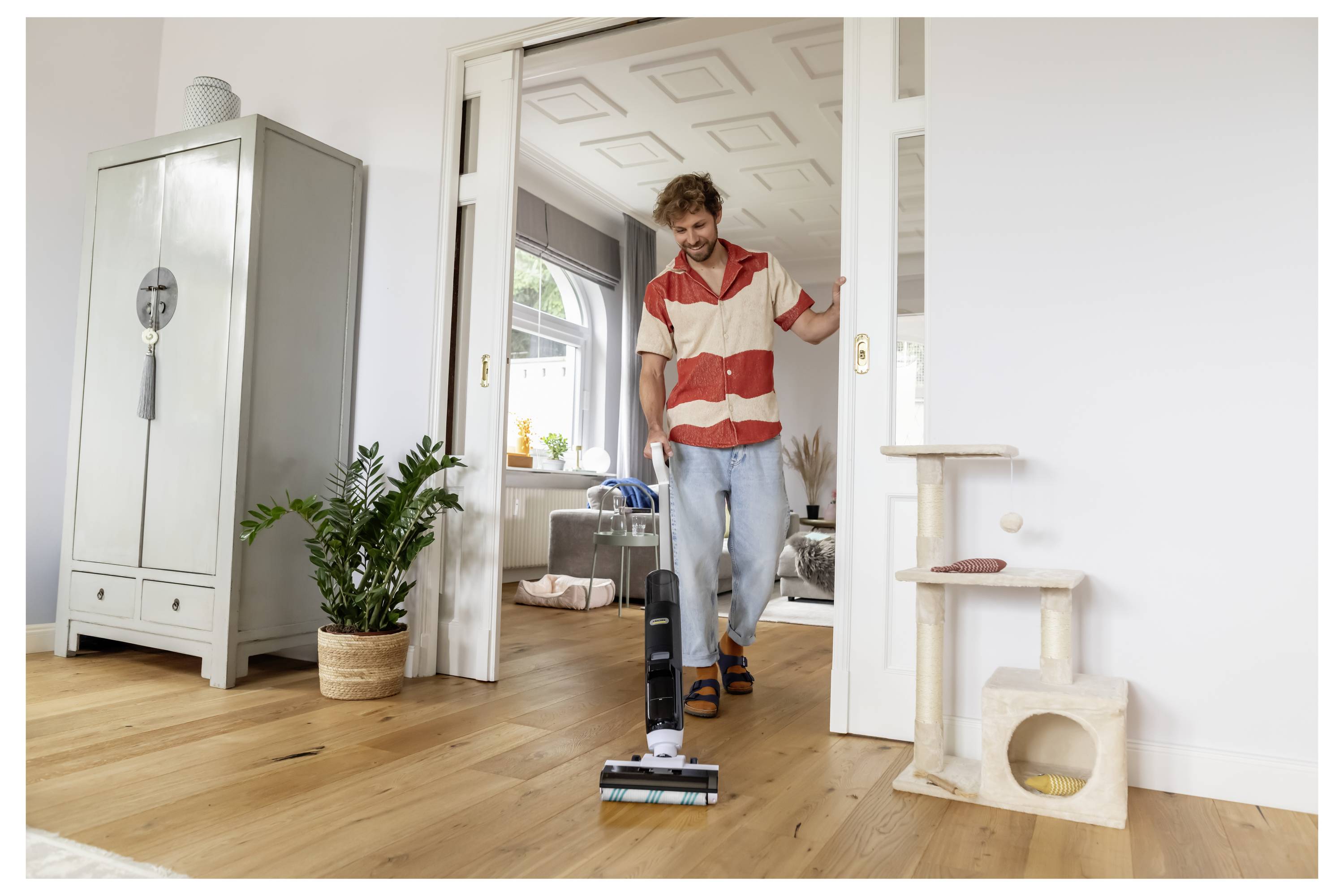 Man vacuuming a hardwood floor in a bright living room, with a cabinet, plant, and cat tree nearby.