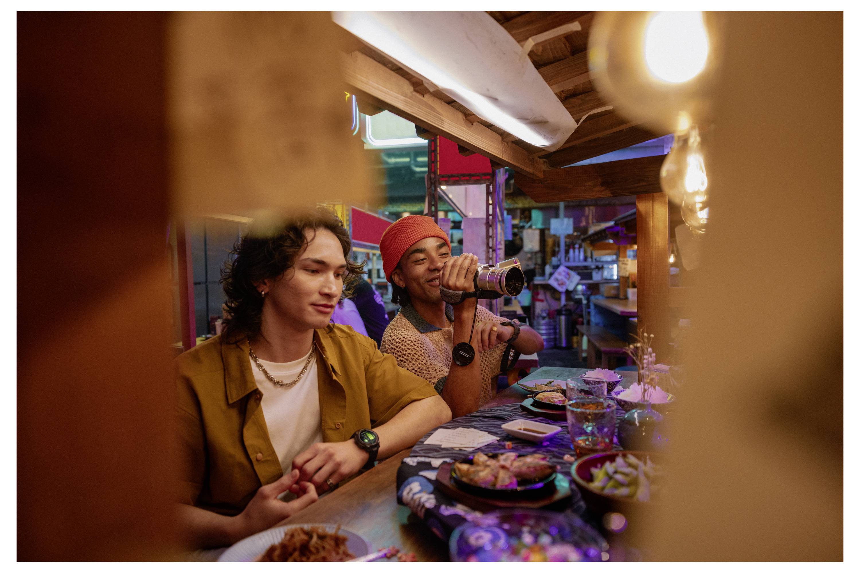 Two people sit at a restaurant table, one drinking from a bottle while food and dishes are spread out in a lively, colorful setting.