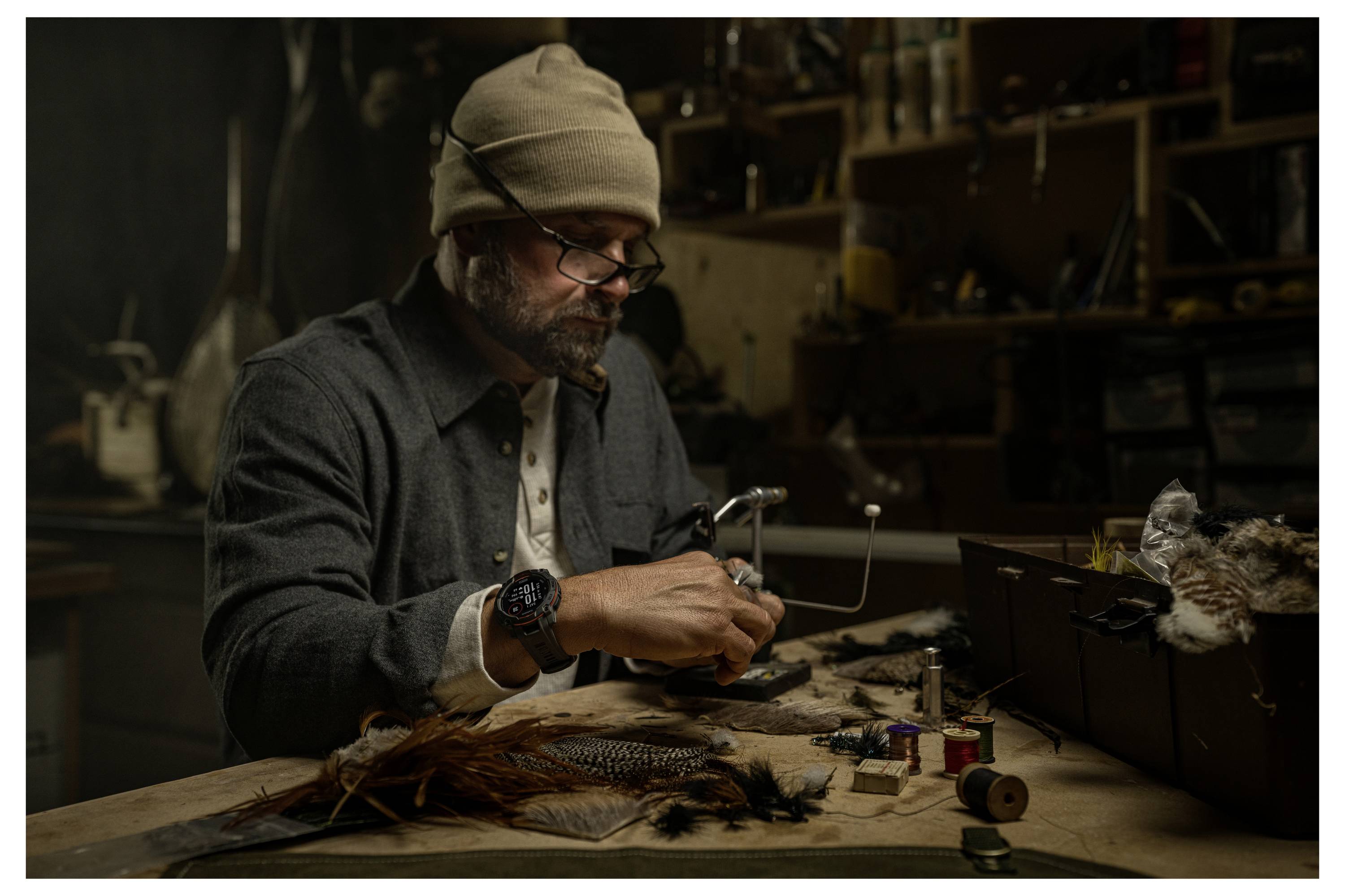 Man in a beanie and glasses works at a cluttered bench, carefully tying a fishing fly or lure with tools and materials spread around.