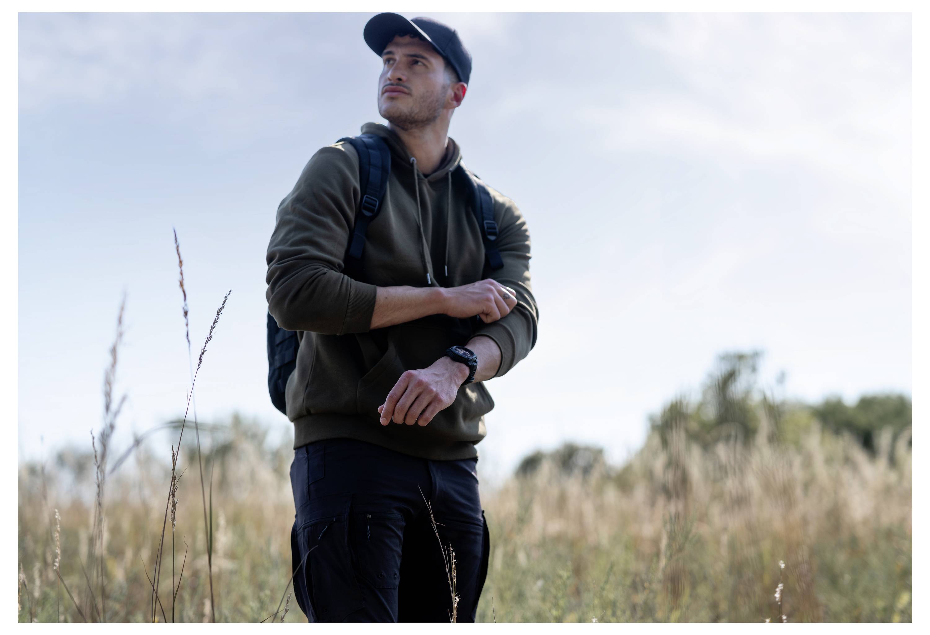 Man wearing a cap and backpack stands in a grassy field, looking into the distance and adjusting his sleeve, suggesting an outdoor hike or exploration.