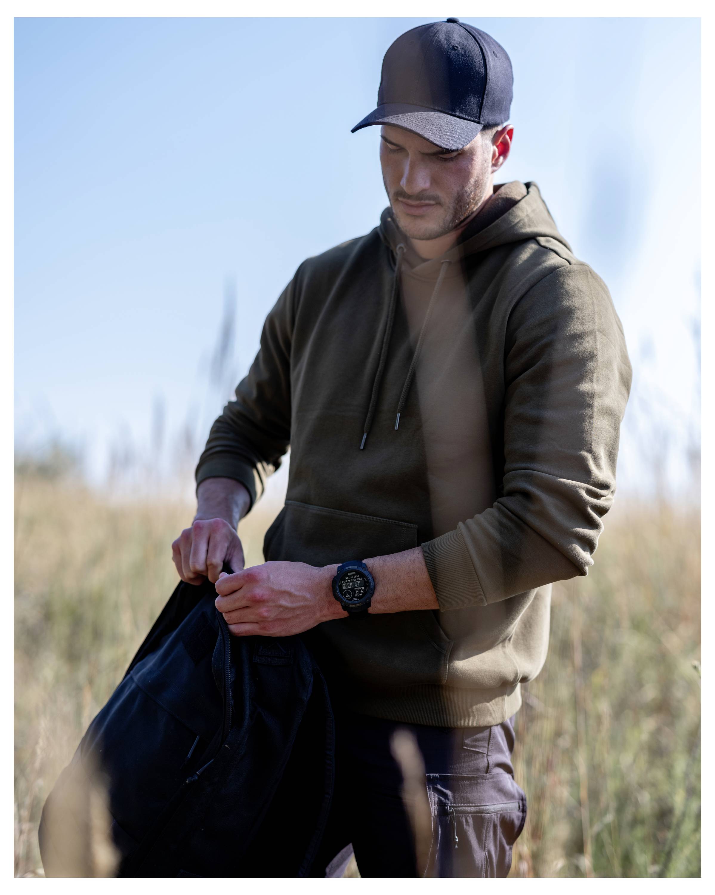 Man outdoors in a field, wearing a cap and hoodie, looking down while handling a black bag or jacket.