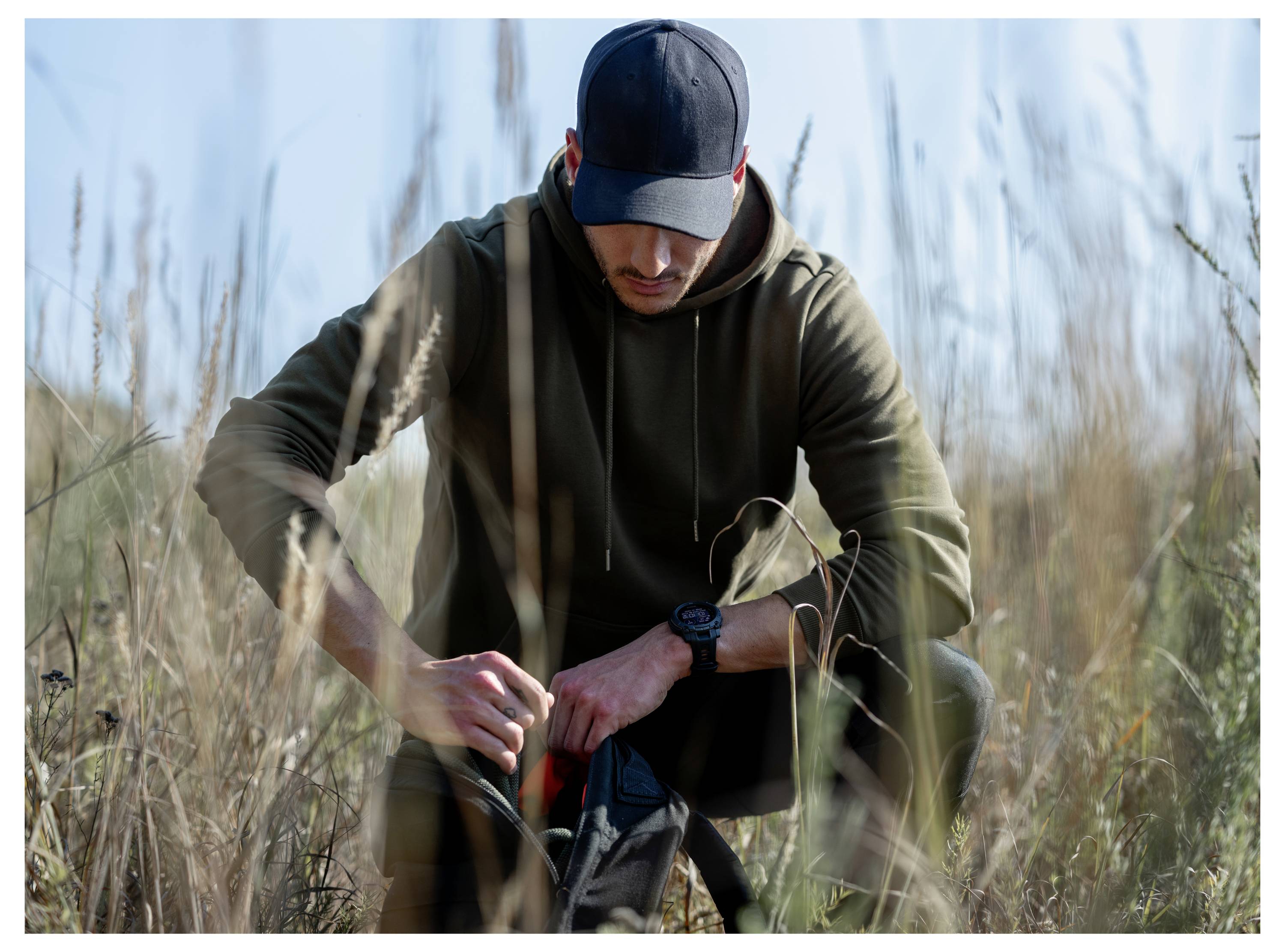A man crouches in tall grass, adjusting or checking gear in a black bag, wearing a cap and hoodie outdoors in daylight.