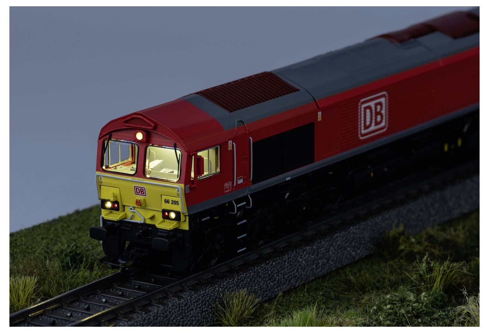 Red DB freight locomotive on tracks at dusk, viewed from the front-left on a grassy embankment.<br>
