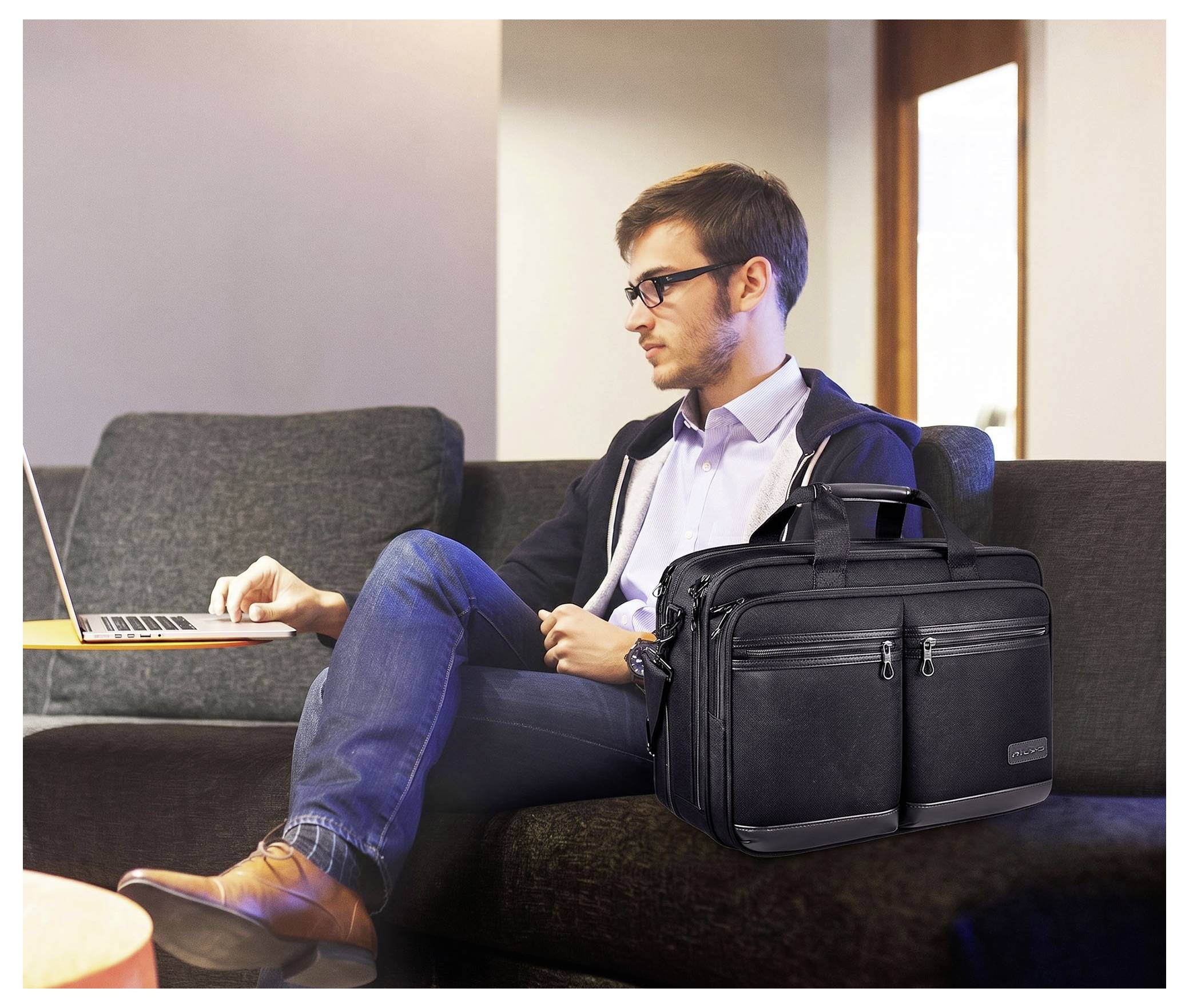 Man seated on a couch using a laptop, with a black briefcase beside him, in a bright living room.<br>