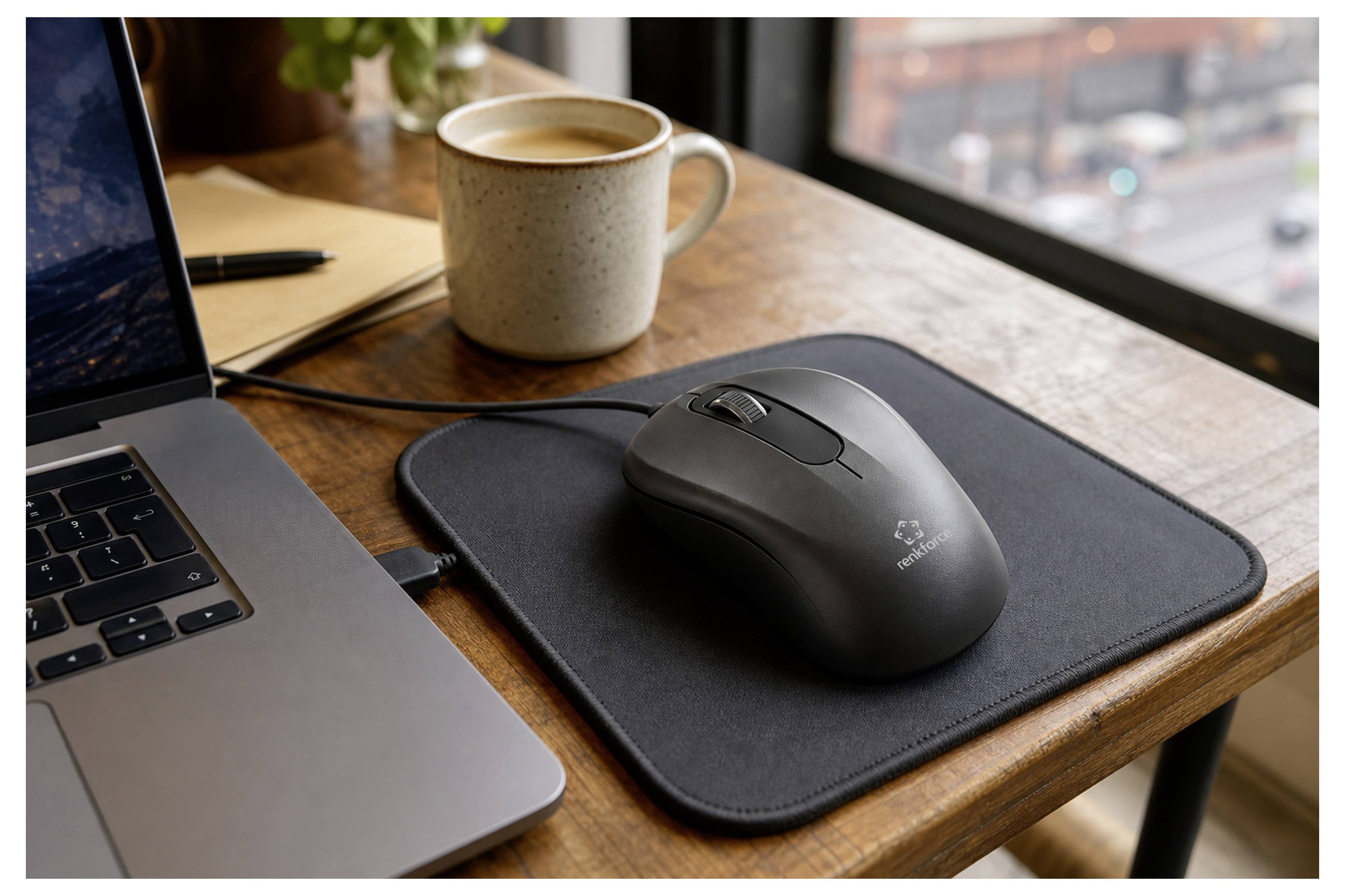 Wireless mouse on a black mouse pad beside a laptop and coffee mug on a wooden desk near a window.