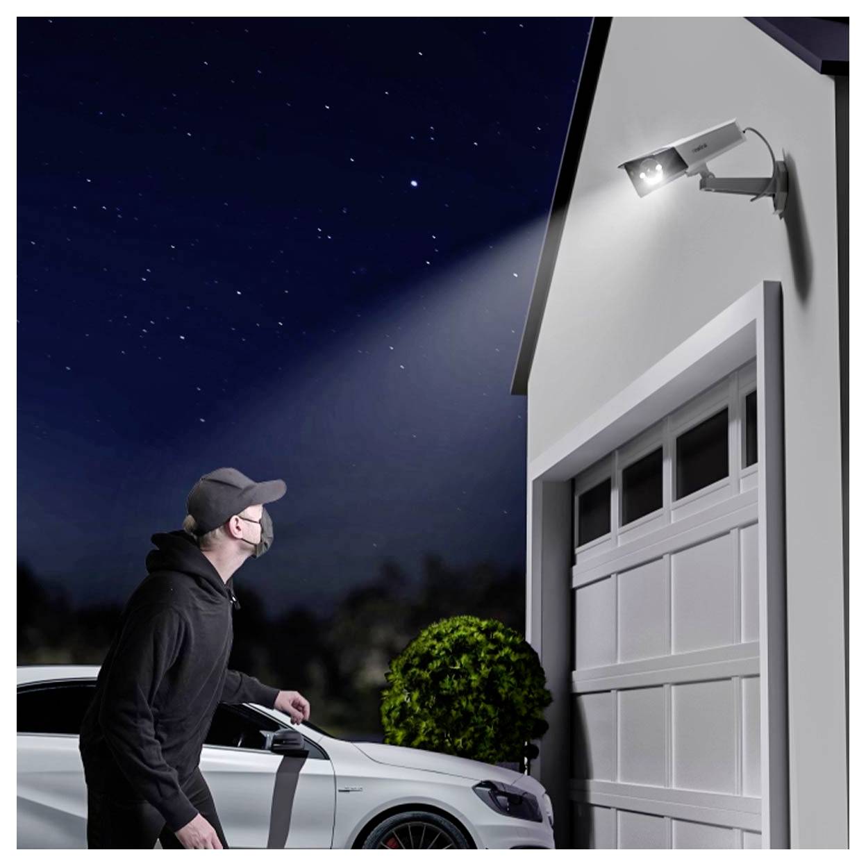 Man at night inspecting a house garage under a bright security light, standing beside a parked car and looking up toward the light fixture.