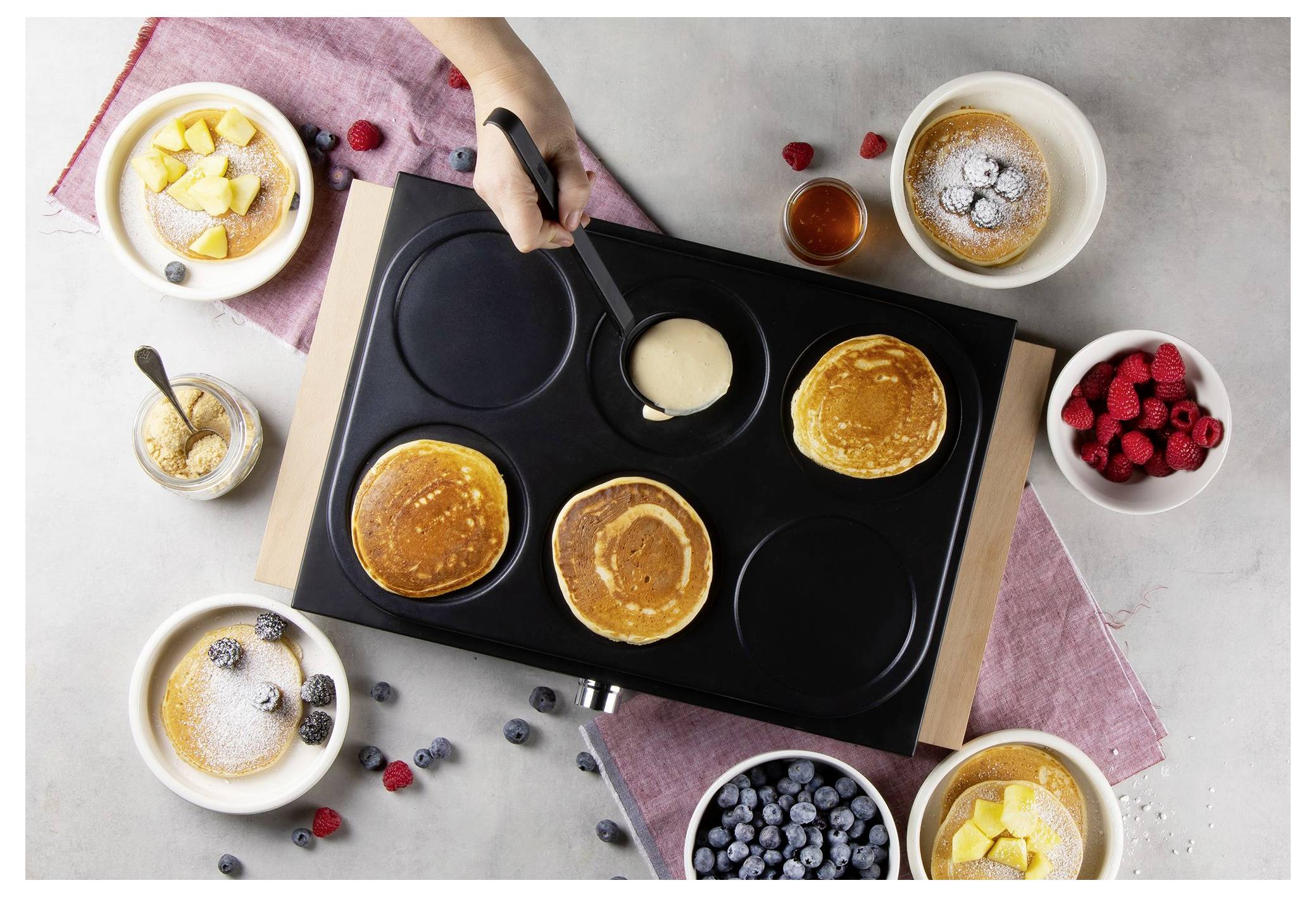 Overhead view of a pancake griddle with batter being poured, surrounded by bowls of fruit, syrup, and finished pancakes.