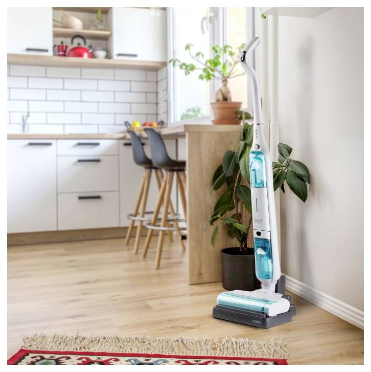 Cordless stick vacuum cleaner stands in a bright kitchen, shown beside a plant and island on a hardwood floor.<br>