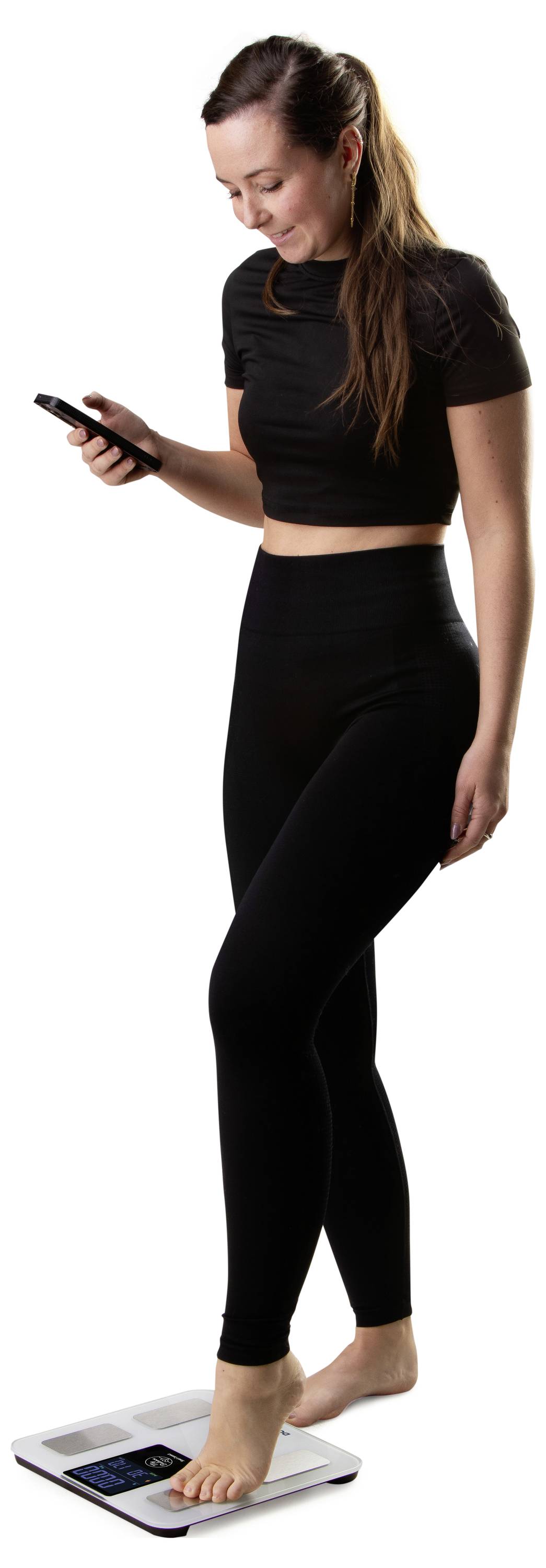 Woman in black workout clothes looking at her phone while standing on a digital bathroom scale.