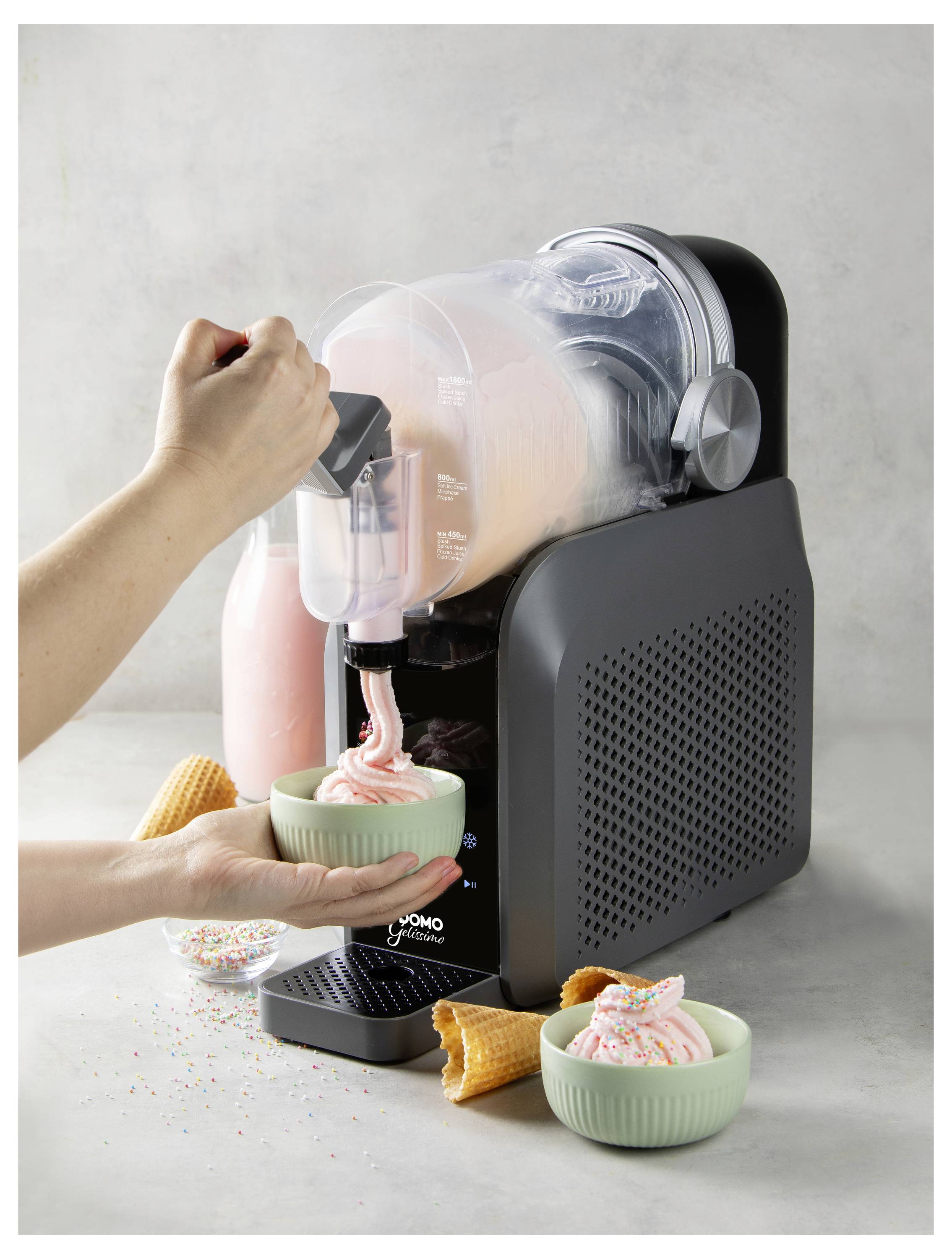 Person dispenses pink soft-serve from a countertop ice cream machine into a bowl, with another topped bowl nearby and waffle cones on the table.