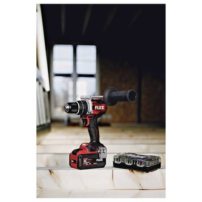 Cordless FLEX power drill with side handle standing on a wood surface beside its battery and case in a workshop-like room.