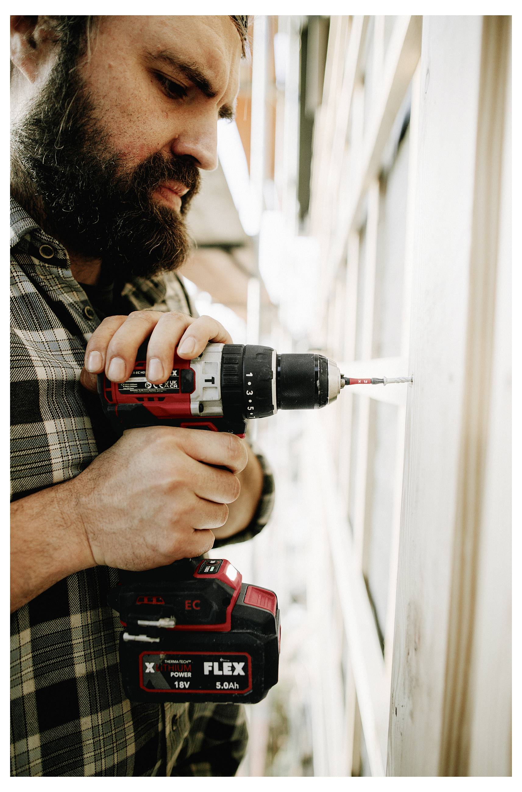 Bearded man drills into a wall with a cordless power drill, focused on the work.