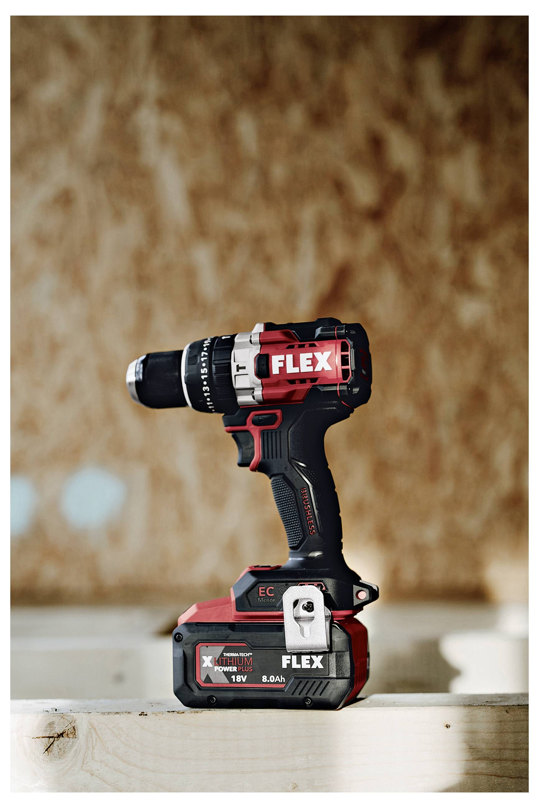 Black-and-red FLEX cordless drill/driver standing upright on a wooden surface against a blurred brown background.
