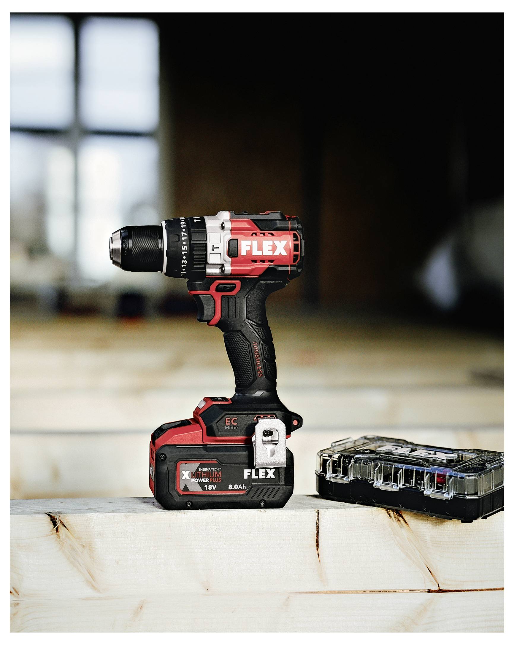 Red and black FLEX cordless drill standing on a wooden surface beside an open battery case, indoors with a blurred background.<br>