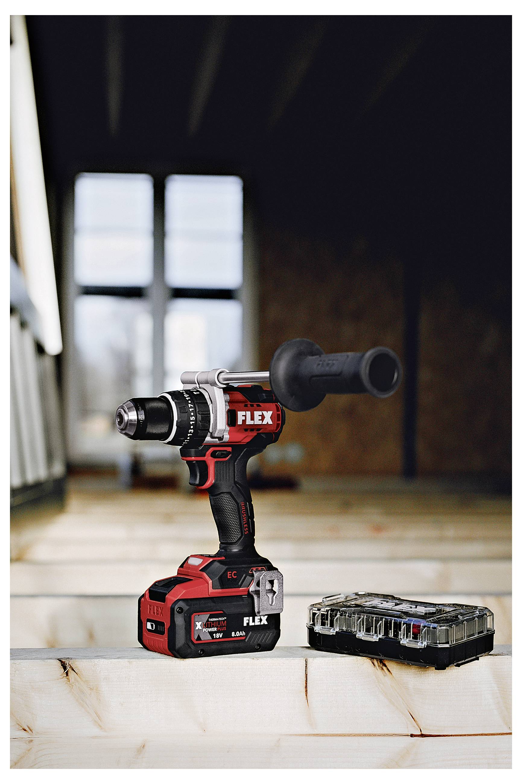 A red-and-black cordless FLEX drill/driver with side handle stands on a wooden surface next to a battery, in a workshop setting.
