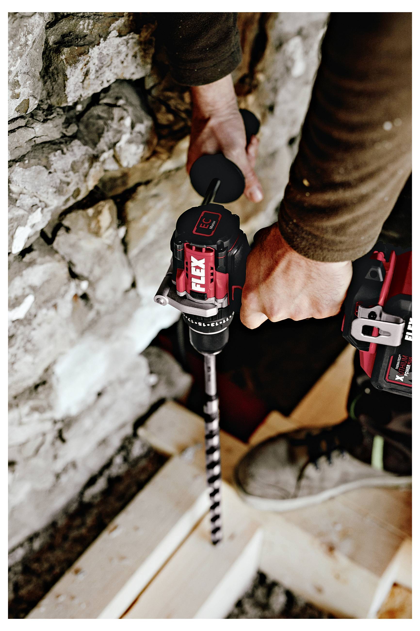 Hands use a cordless drill to bore a hole into wood beside a stone wall during construction work.