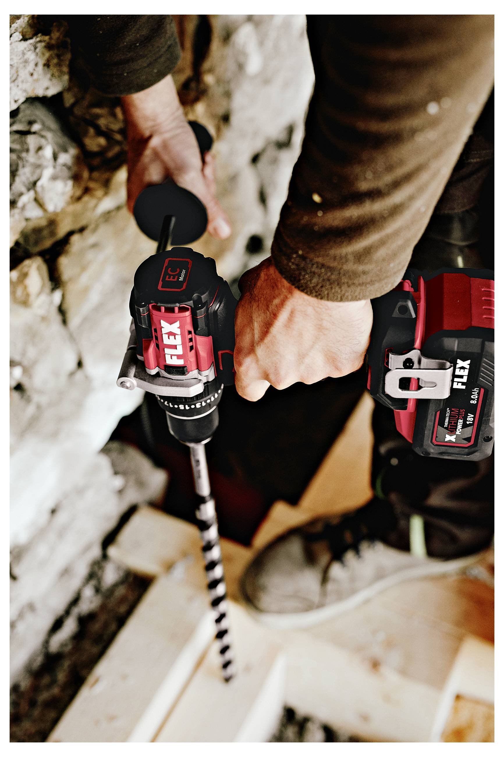 Person using a cordless power drill with a long auger bit to bore into a wall or floor, likely during construction or renovation.