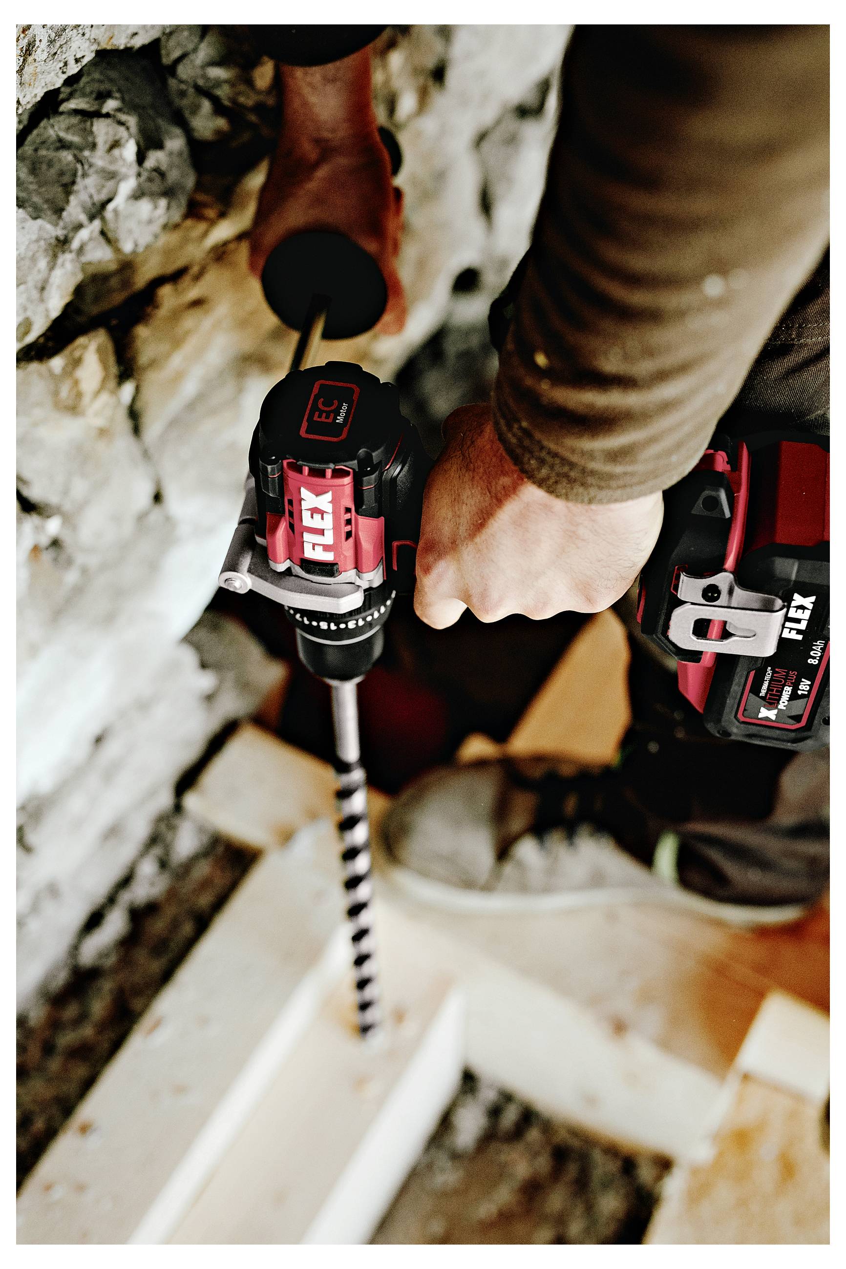 Person using a cordless FLEX drill with a masonry bit to drill into a stone wall or floor at a construction site.
