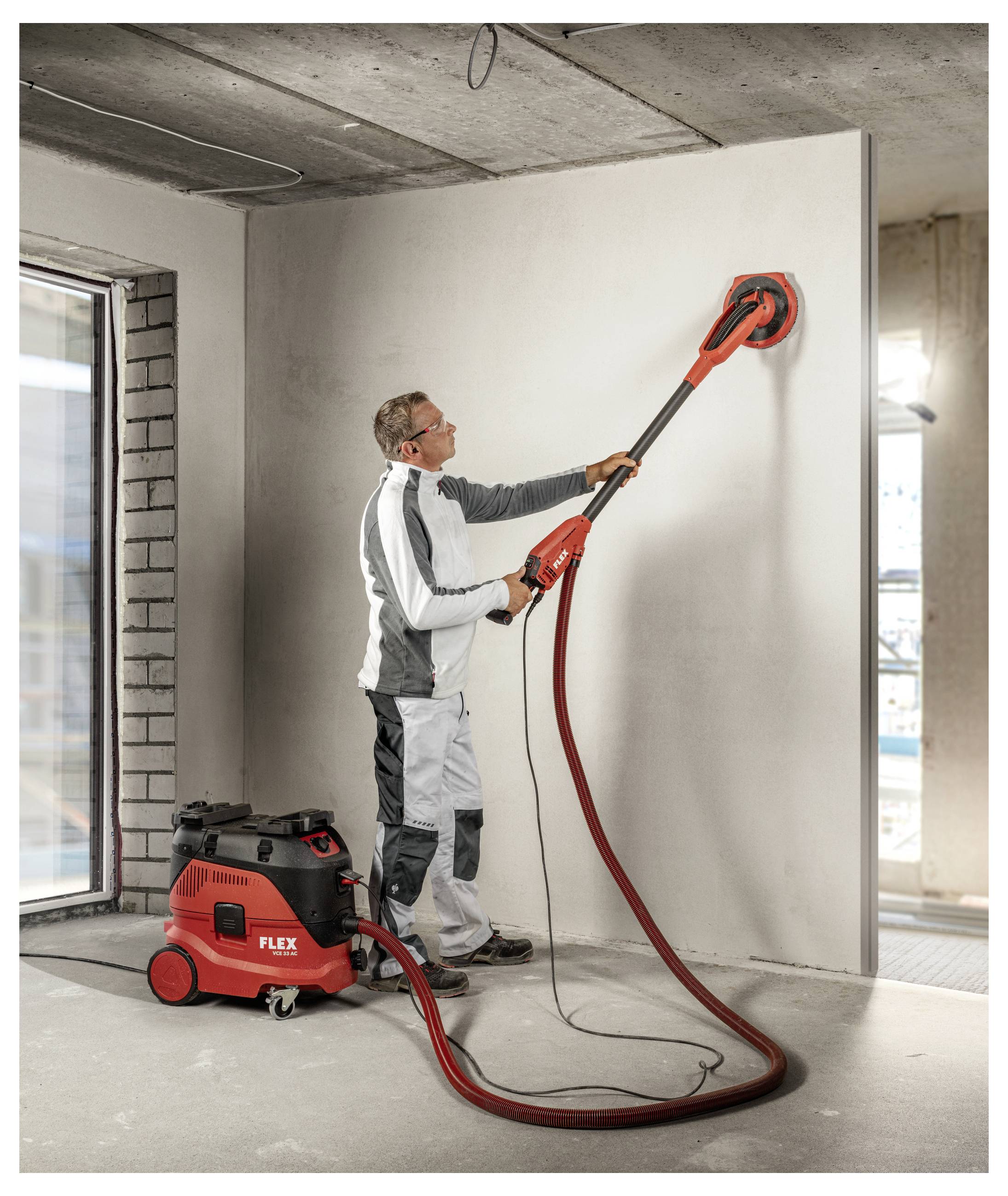 Worker using a red long-handled drywall sander to smooth an interior wall, with a vacuum unit on the floor and exposed construction setting.