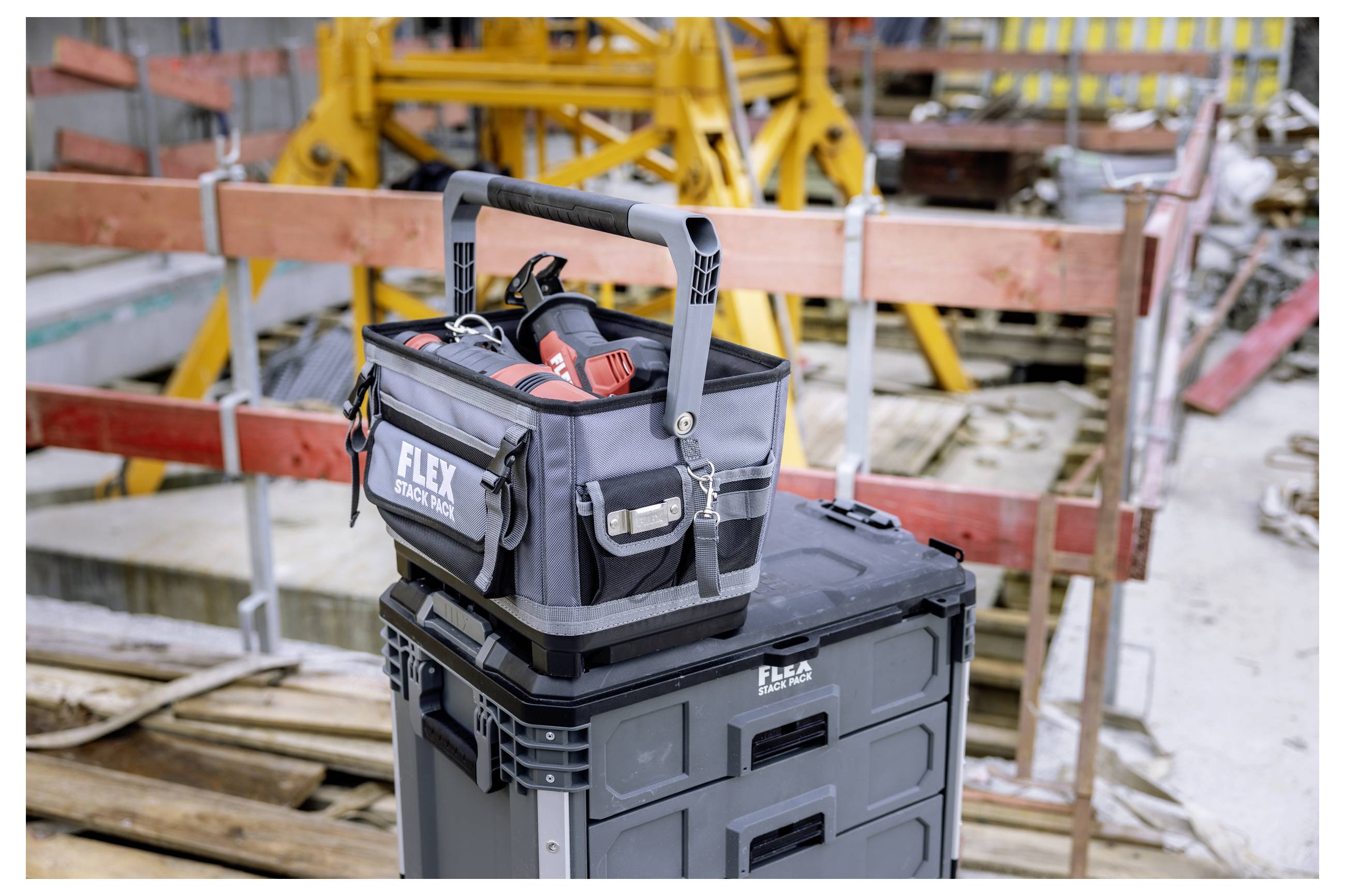 Tool bag with power tools sits on a black storage case at a construction site, surrounded by scaffolding and wooden barriers.