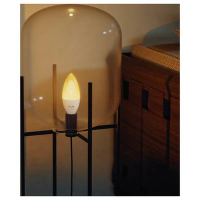 Lit Philips Hue-style candle bulb inside a glass table lamp, beside a wooden cabinet in warm indoor light.