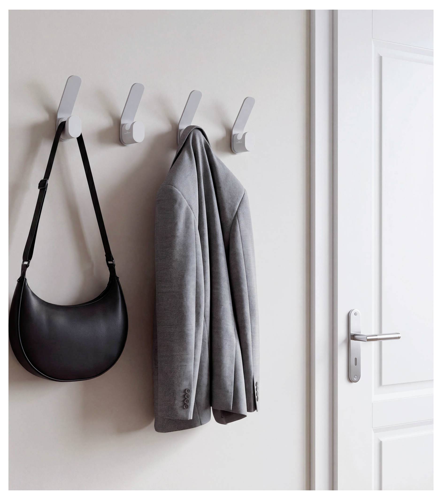 Wall-mounted hooks hold a black shoulder bag and gray jacket beside a white door.
