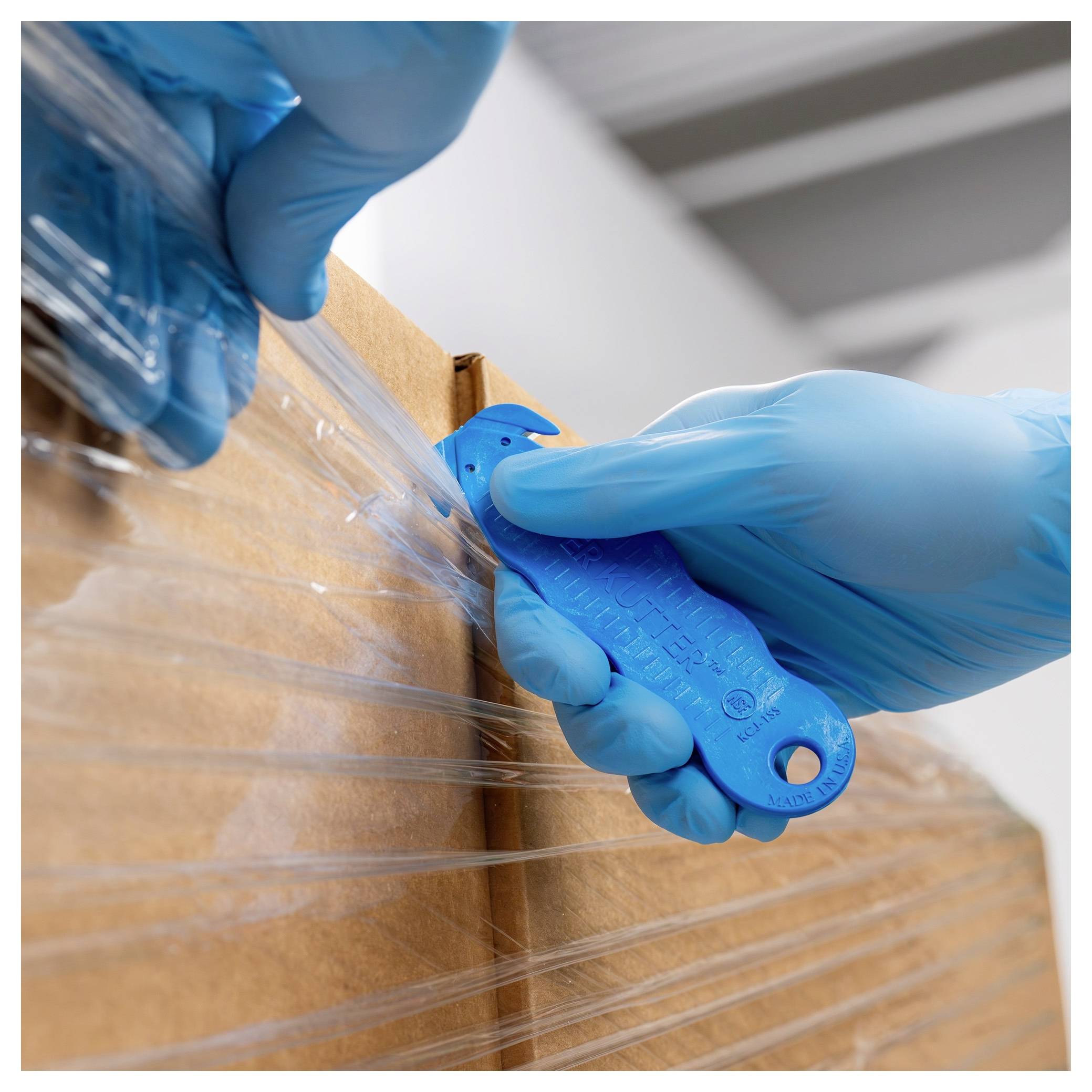 Blue-gloved hands use a blue utility knife to cut clear plastic wrap from a cardboard box.