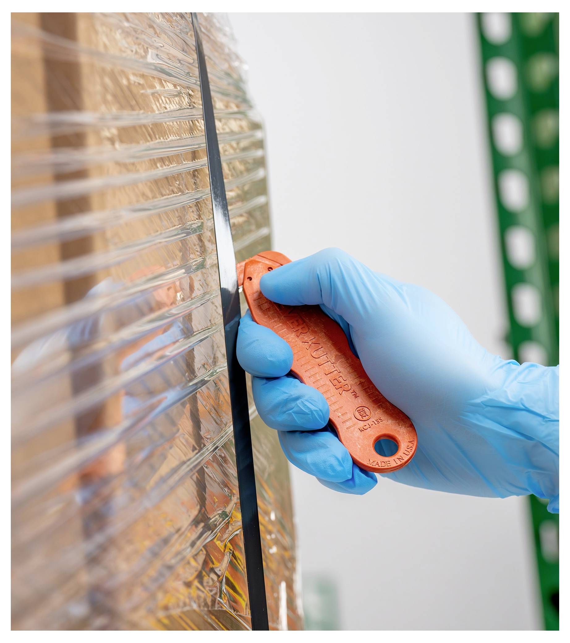 Gloved hand using an orange utility knife to cut a black seam on reflective insulating foil or packaging material.<br>