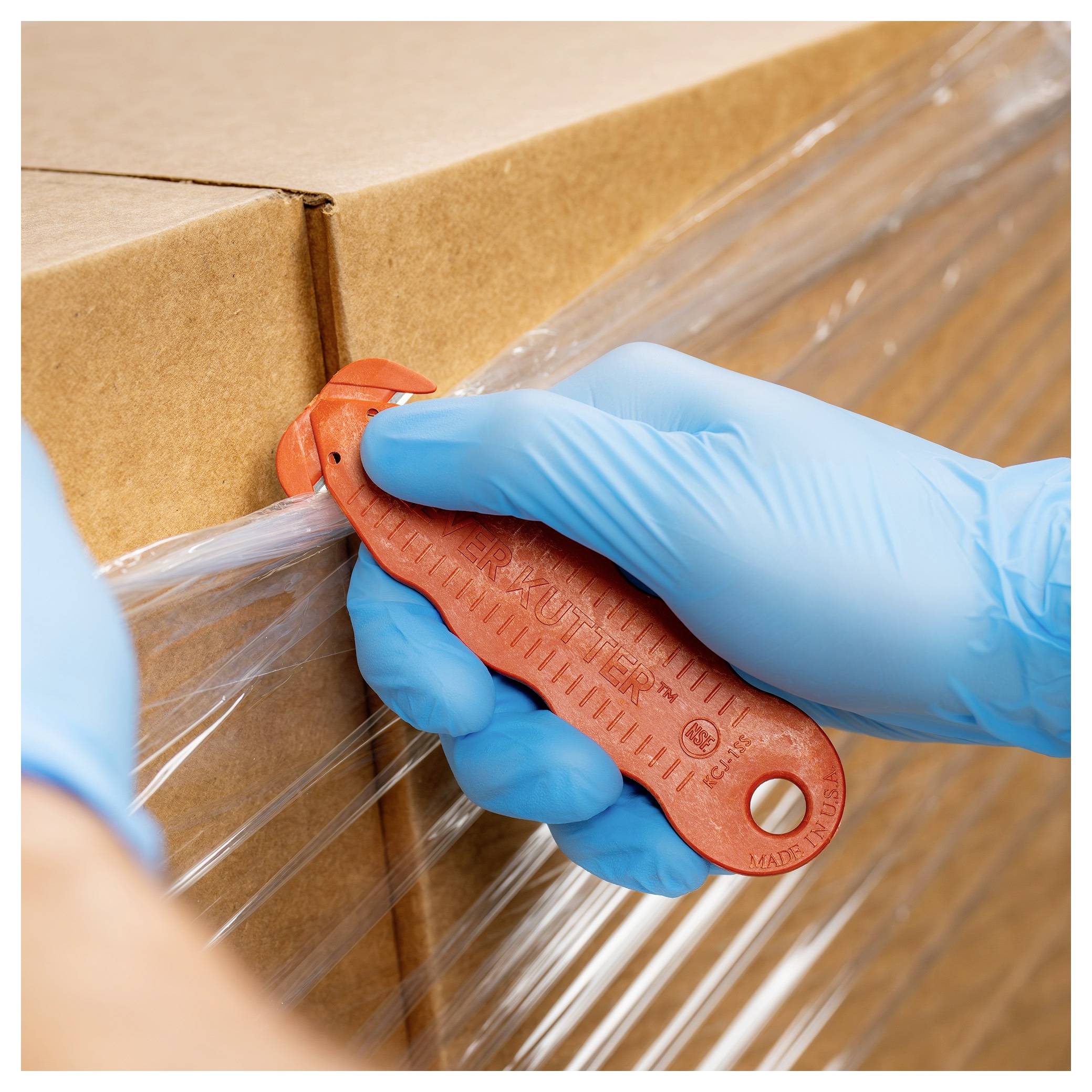 Gloved hands use a red box cutter to slice clear plastic wrap around a cardboard shipping box.