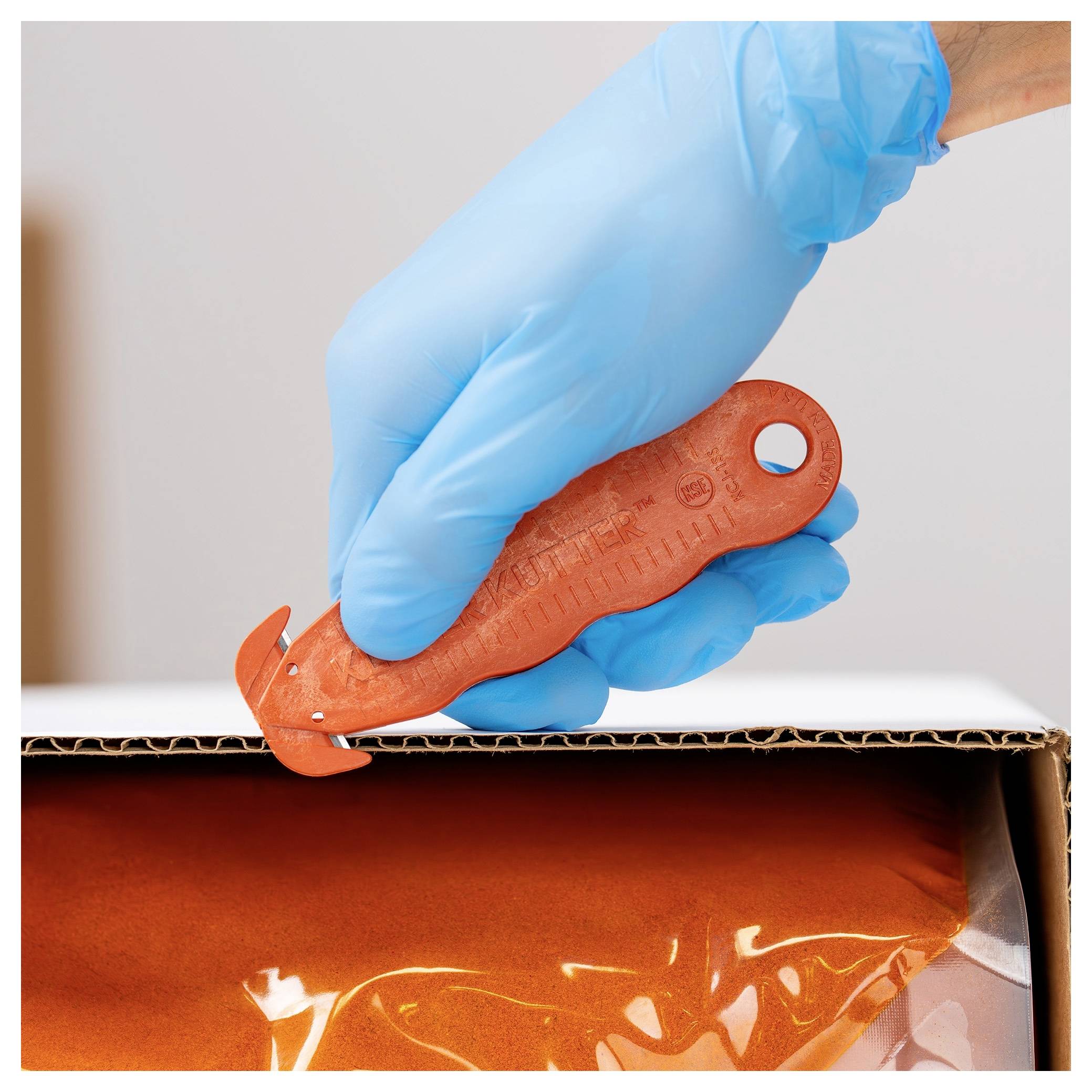 Gloved hand uses an orange box cutter to open a cardboard box containing orange packaging material.
