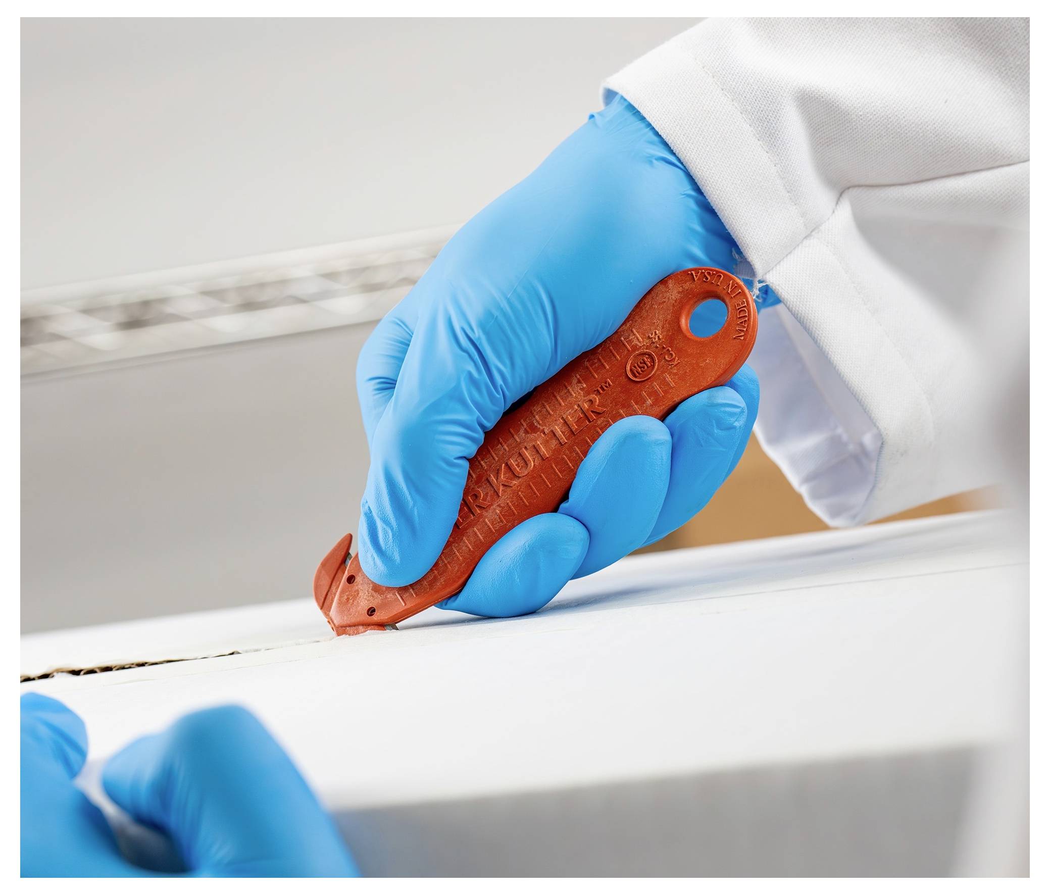 Blue-gloved technician uses a red utility knife to cut along the edge of a white surface, likely opening or trimming packaging.