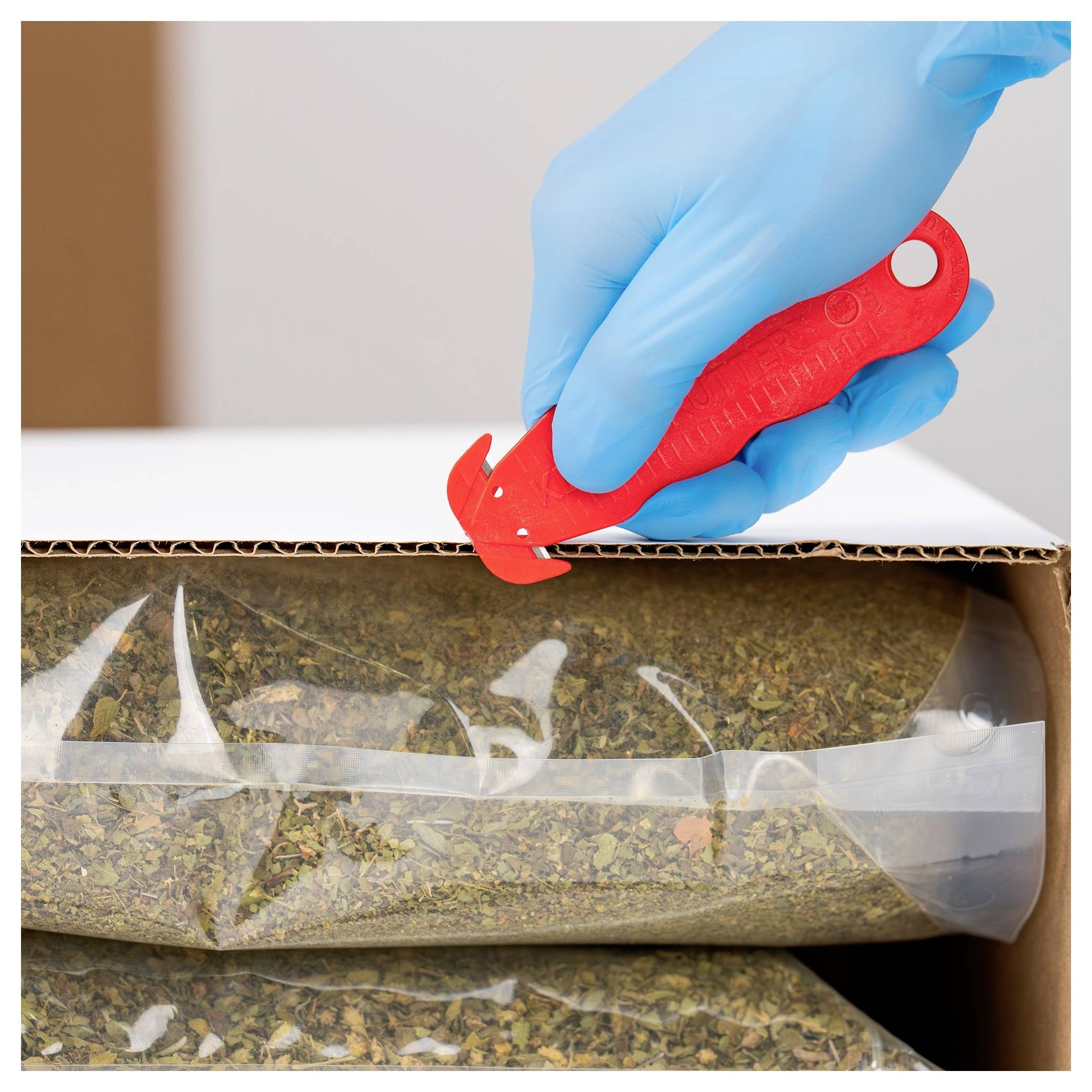 Gloved hand using a red box cutter to open a cardboard package containing a sealed plastic bag of dried herbs or tea leaves.